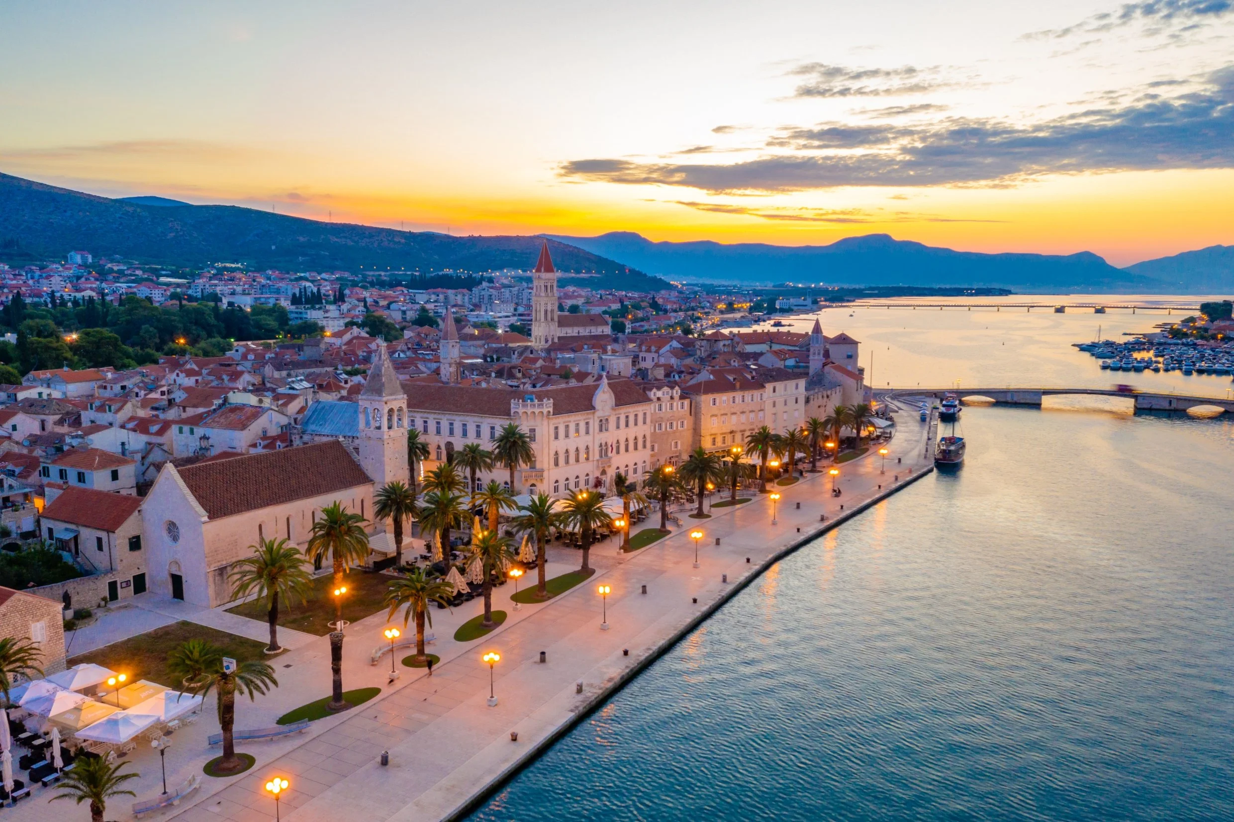 Promenade in Trogir, next to buildings and palm trees with a sunset in the mountains behind the city