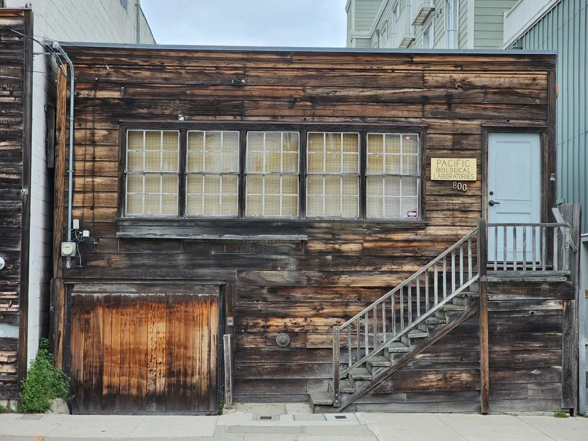A squat building - Pacific Biological Laboratories - made of horizontally laid timber with steps up to a door on the first floor.