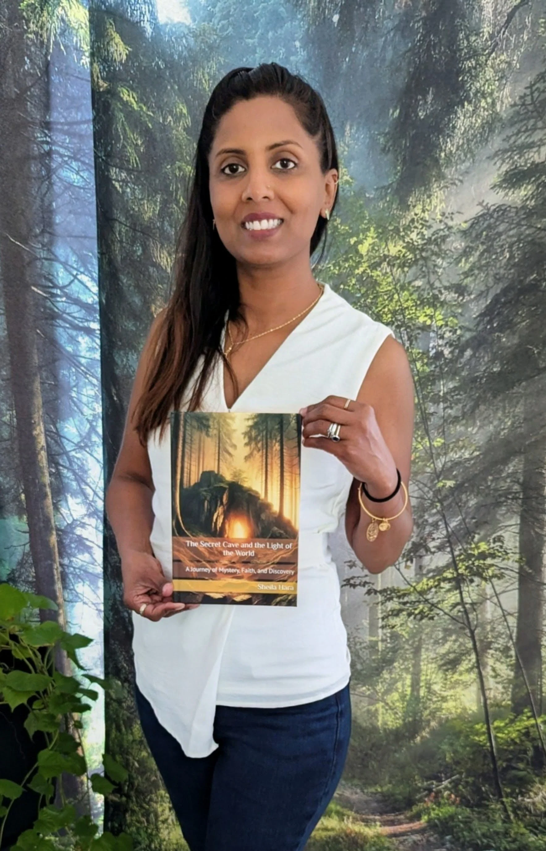 A woman in a white sleeveless top and dark jeans standing in front of a forest-themed background, holding a book titled 'The Secret Cave and the Light of the World' by Sheila Flara.