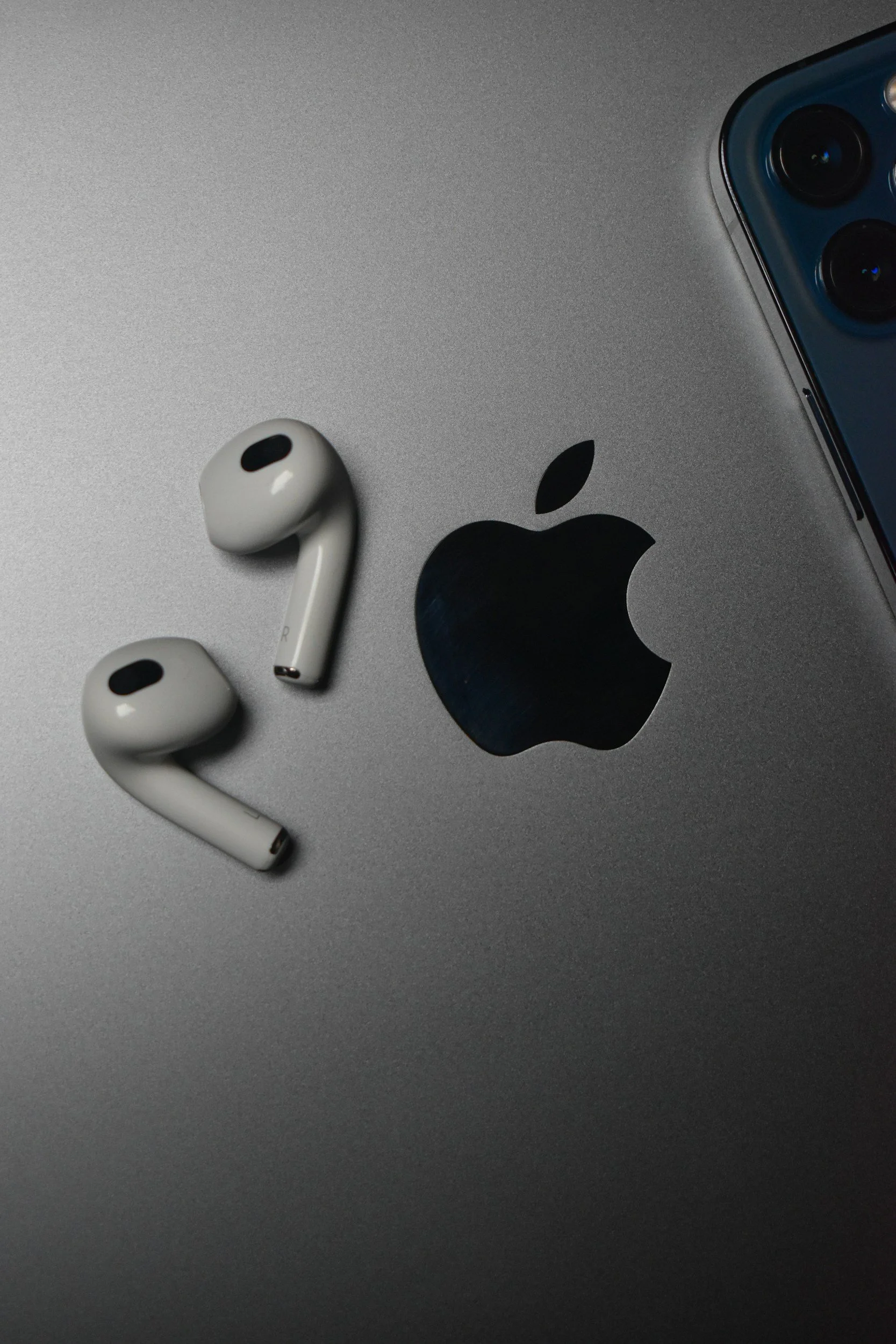 Close-up of a silver Apple laptop with the Apple logo, a pair of white wireless earbuds, and a blue iPhone 13 arranged on top.