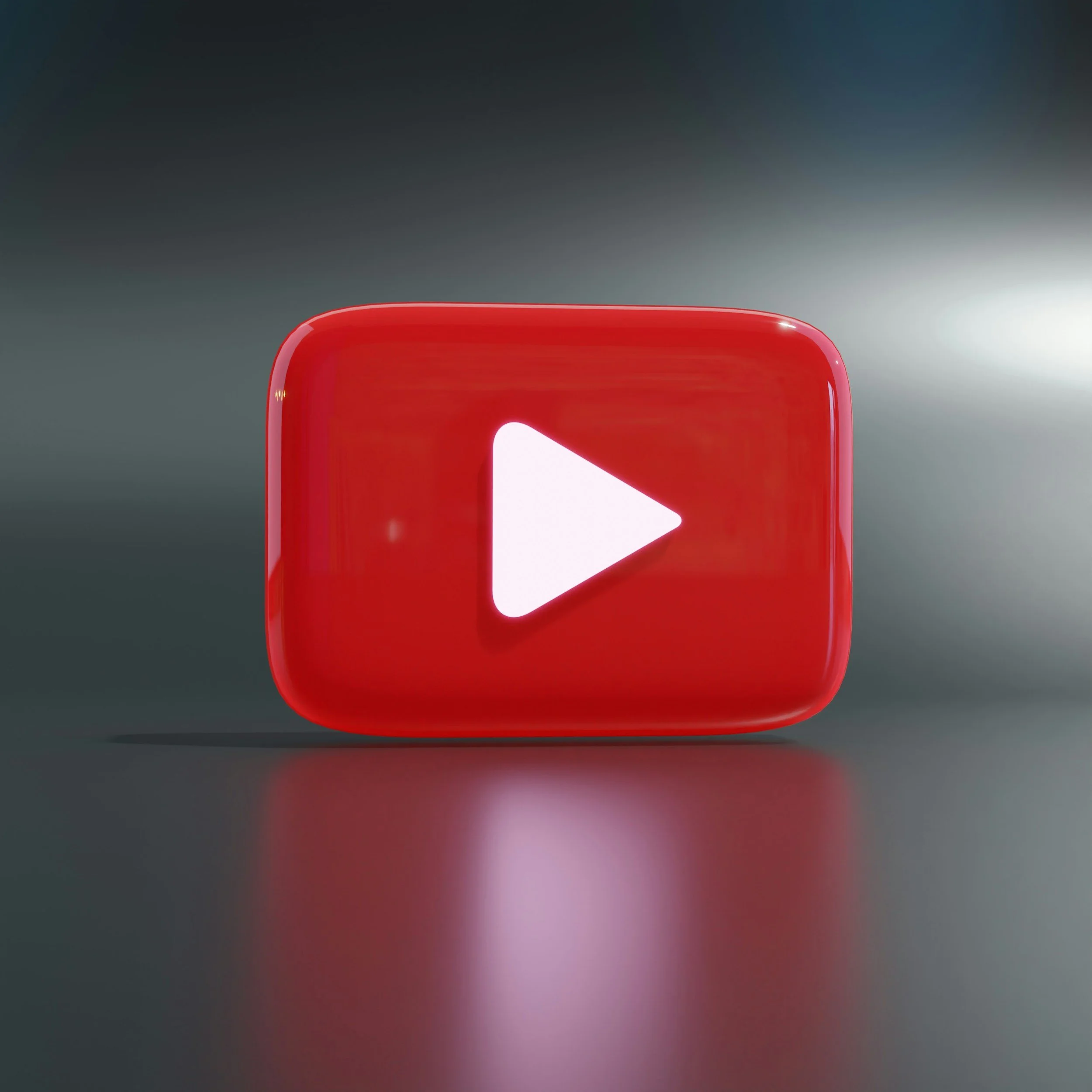 Red YouTube play button icon illuminated with reflection on grey surface.
