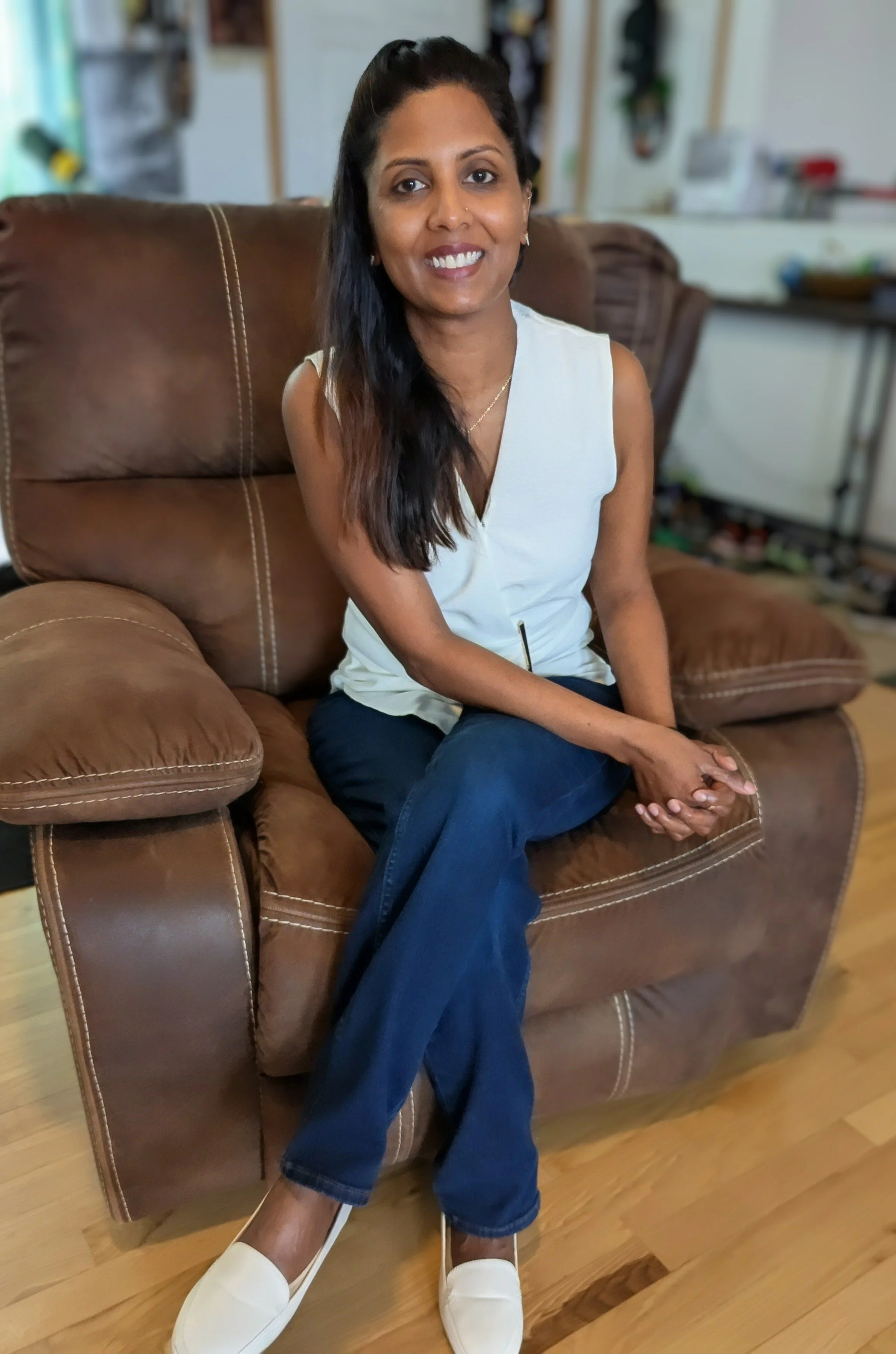 A woman sitting on a brown leather couch in a living room, smiling at the camera.