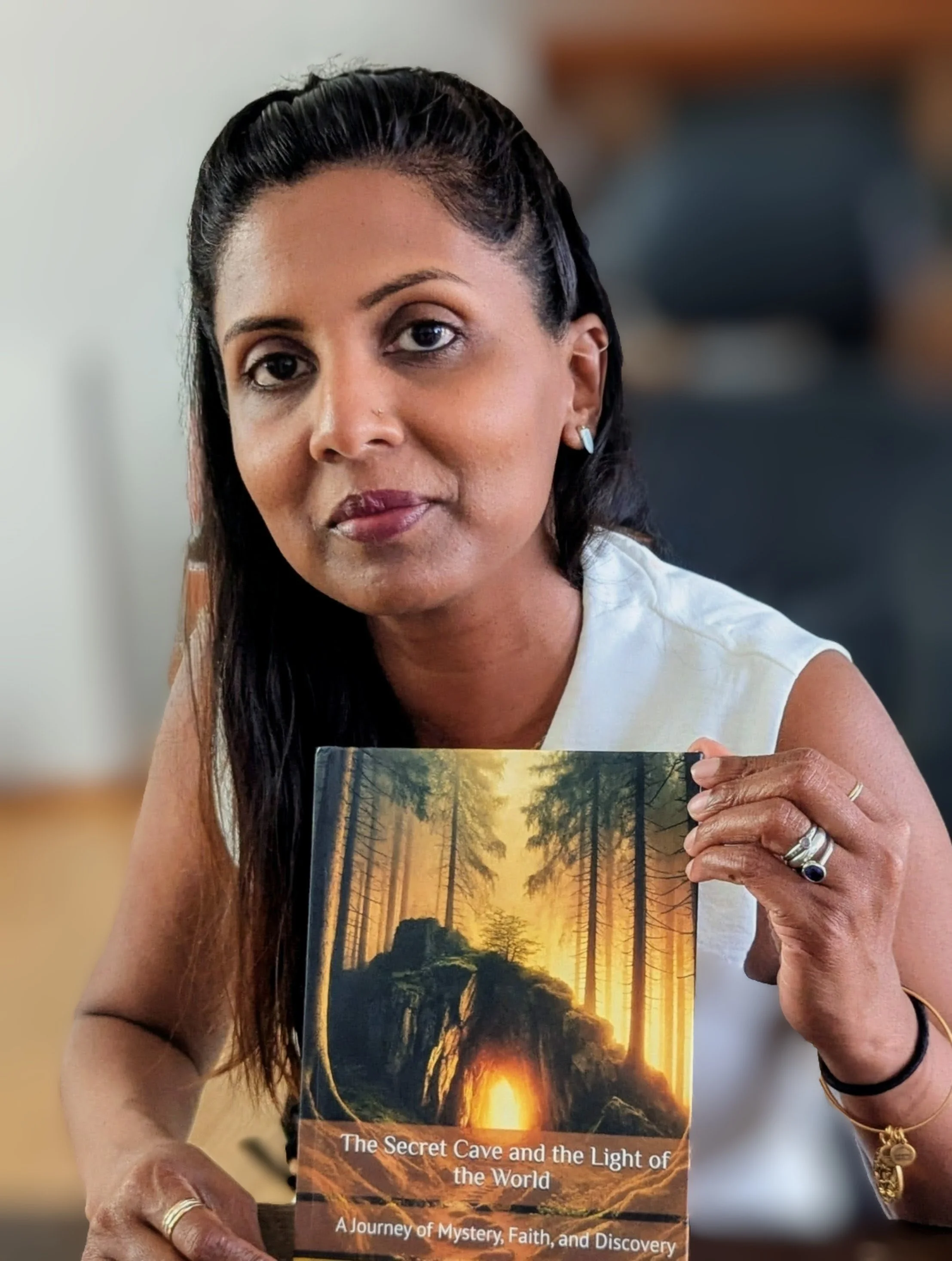 A woman with dark hair and earrings holds a book titled 'The Secret Cave and the Light of the World' showing a cave opening surrounded by trees with a glowing light outside.
