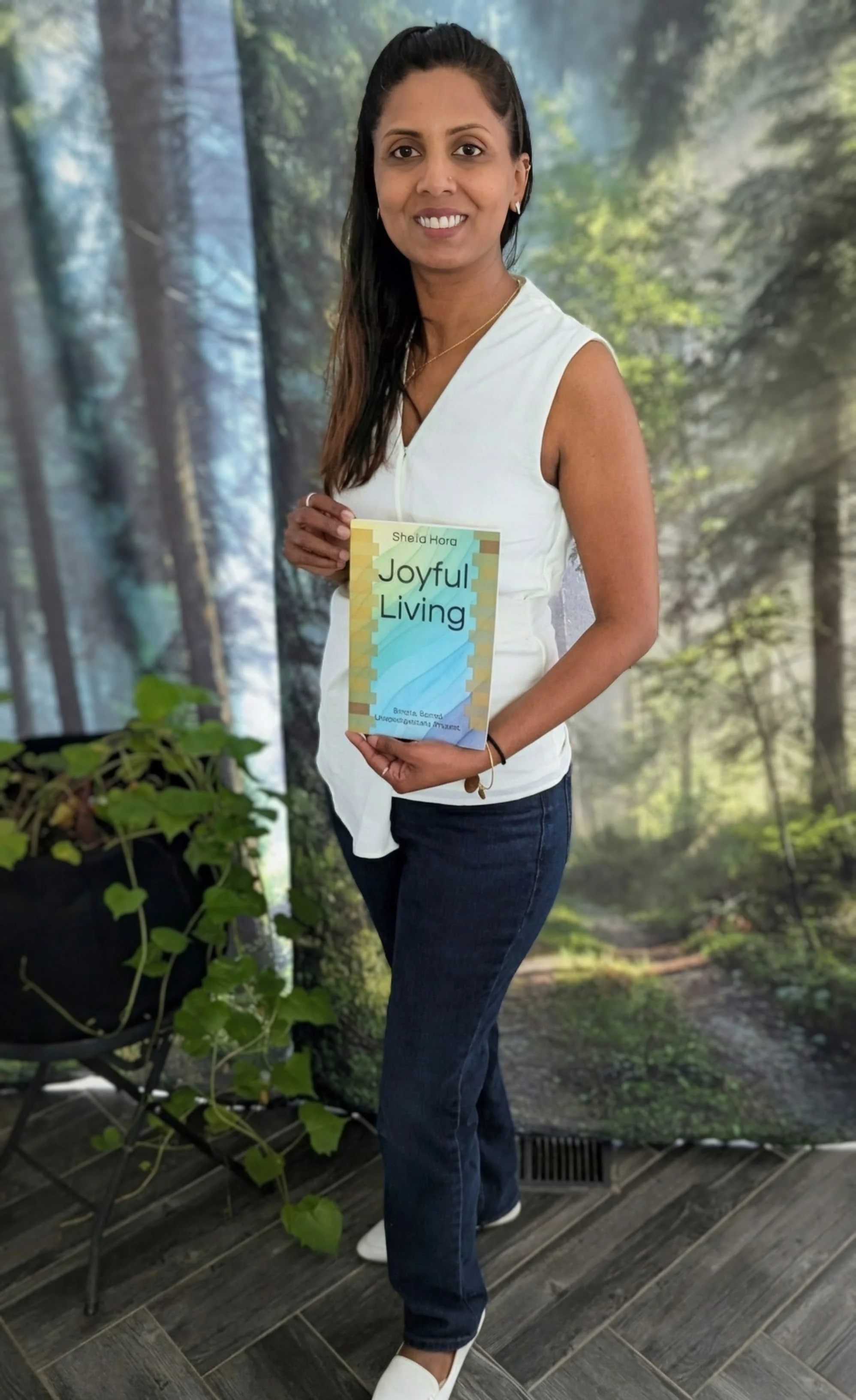 A woman with dark hair, wearing a white sleeveless top and dark jeans, stands in front of a forest-themed backdrop, holding a book titled 'Joyful Living' by Sheila Hora.
