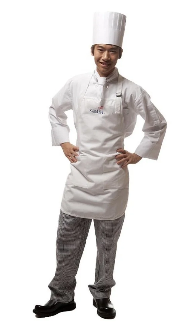 A culinary student models a white chef’s uniform with a tall hat.