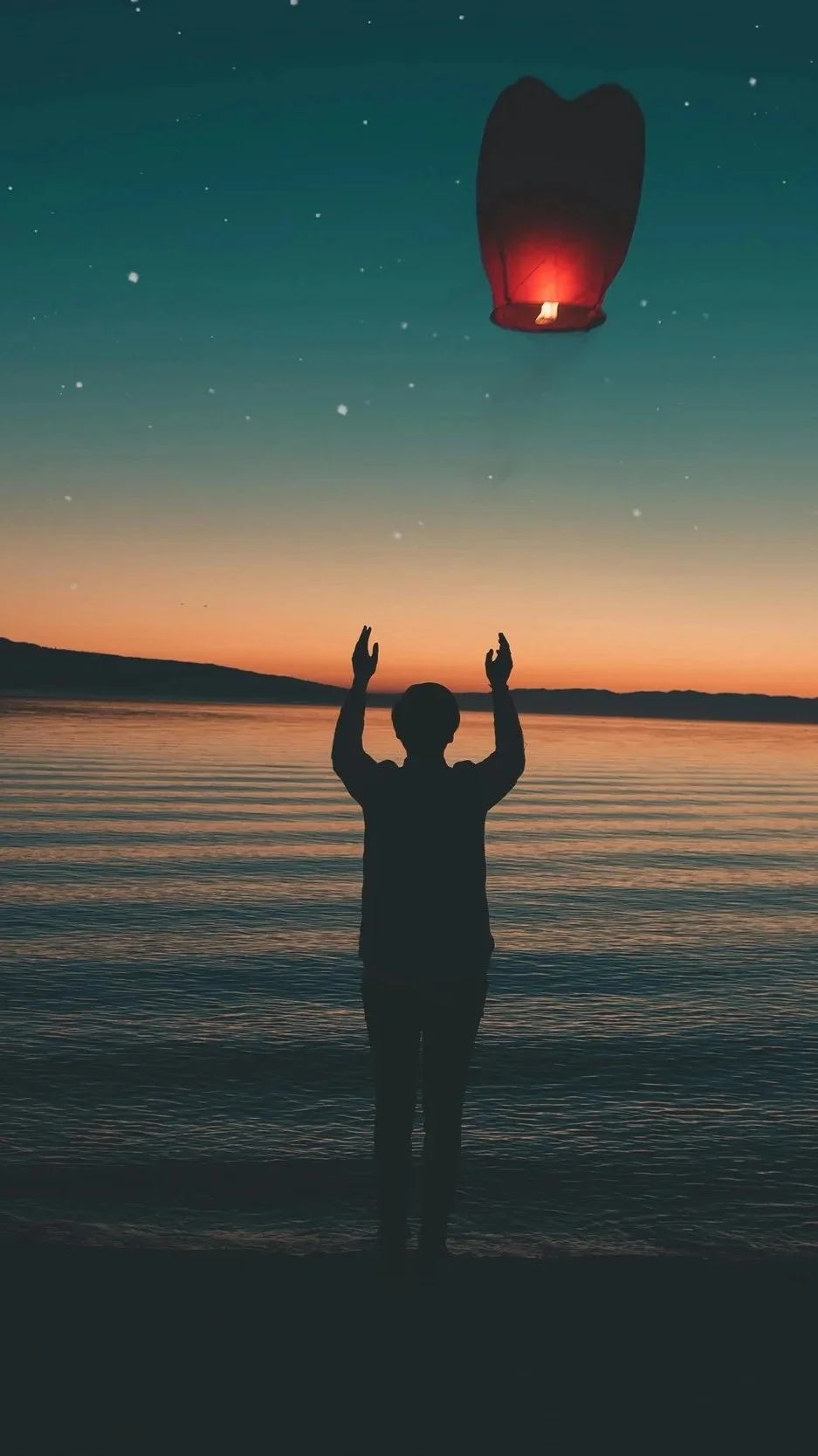 A person standing by the water during sunset, releasing a sky lantern into the sky.