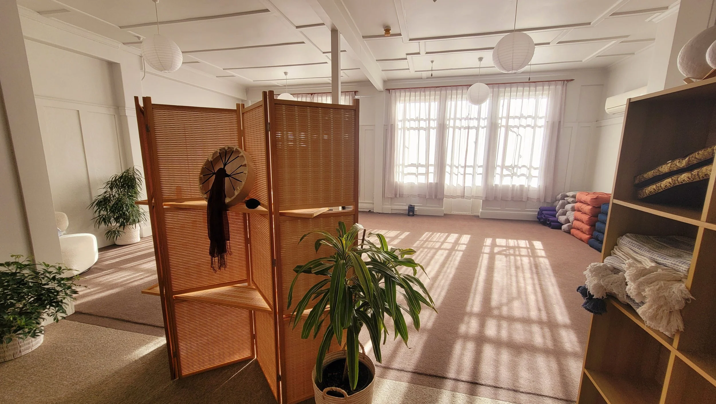 Bright yoga or meditation studio with large windows, potted plants, and neatly stacked mats and blankets along the walls, divided by a wooden partition.