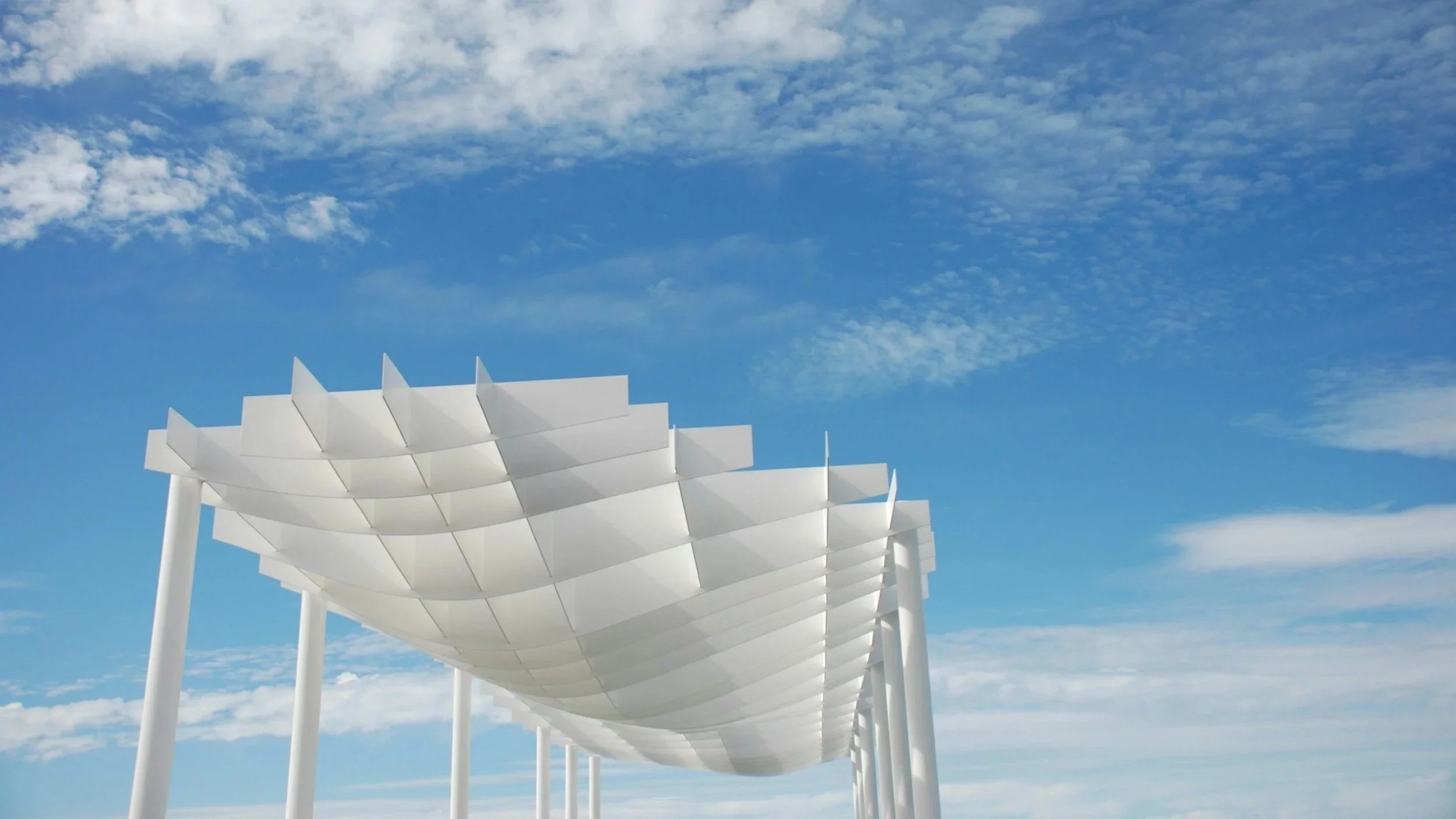 Modern white architectural structure with curved panels supported by tall white pillars against a partly cloudy blue sky.