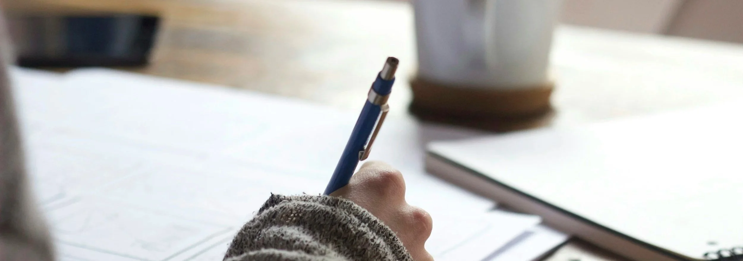 A person holding a blue pen writing on paper with a gray knitted sleeve visible, a white cup and a closed notebook on a table.