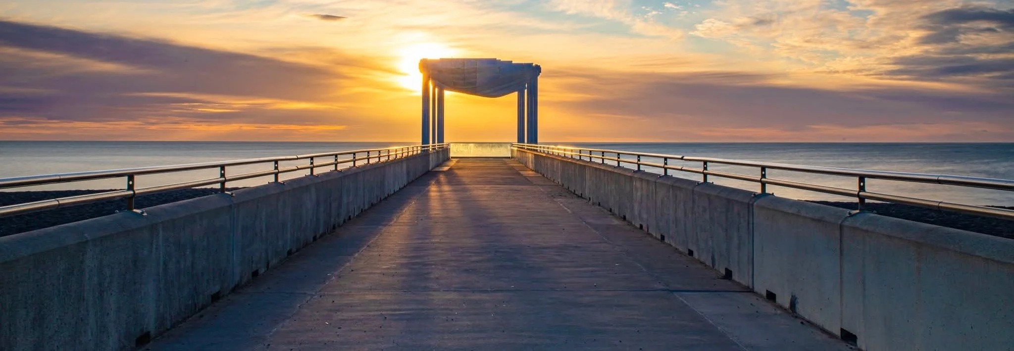 A concrete pier extending into the ocean at sunset with a unique structure at the end, the sky with orange and pink hues and the sun partially obscured.