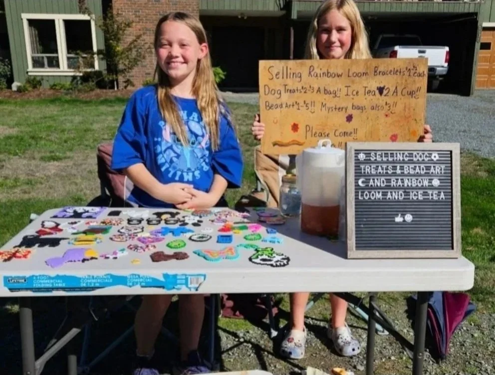 Two young girls running a lemonade stand outdoors with colorful bracelets on the table. One girl is holding a sign advertising Rainbow Loom bracelets, dog treats, ice tea, and beads. The other girl is sitting next to the table, smiling.