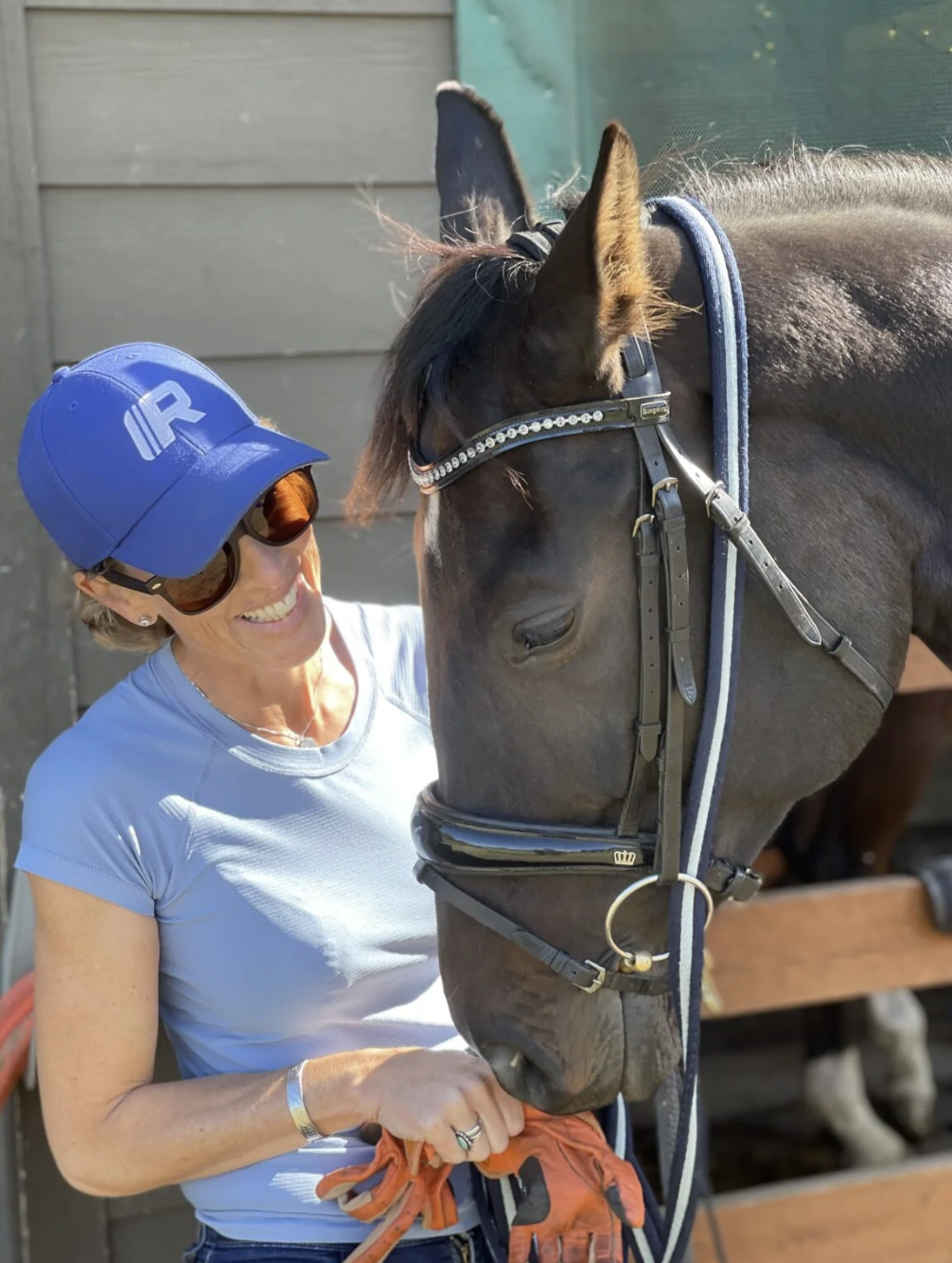 A woman in a blue cap and sunglasses smiling while petting a dark brown horse with a bridle inside a stable.
