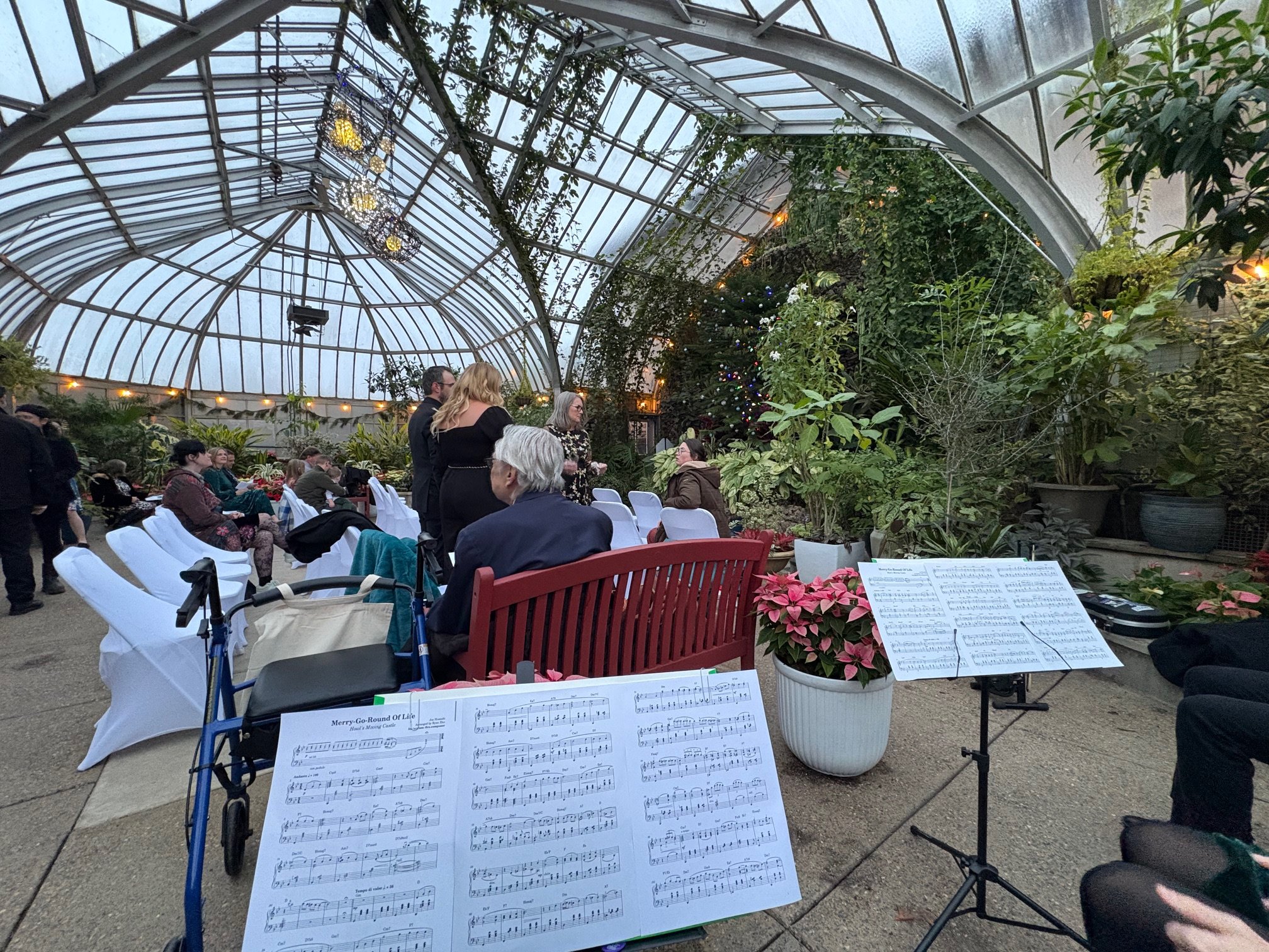 In the foreground are several stands set up with sheet music on them; in the background, a beautiful, leafy conservatory. People mill about.