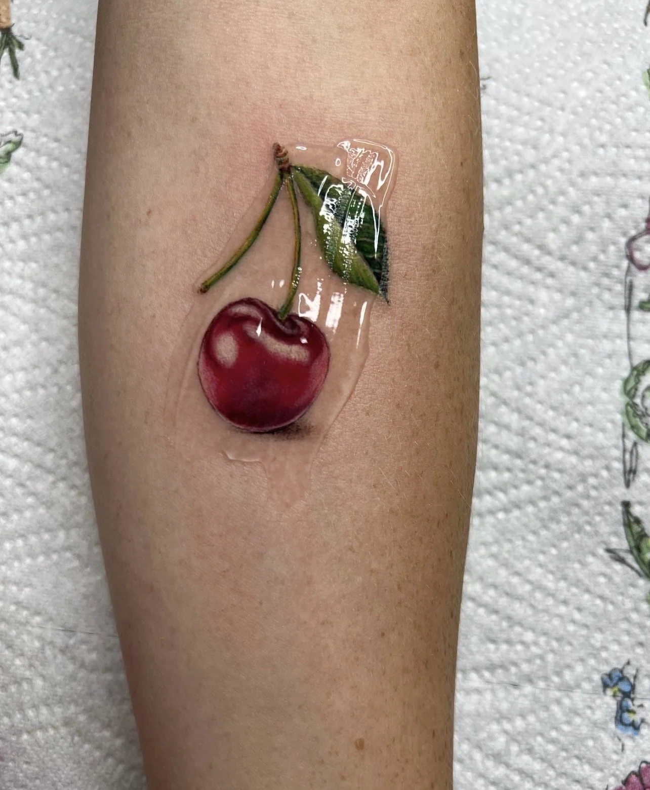 A tattoo of a cherry with a leaf and stem on a person's arm, covered with a transparent protective film.