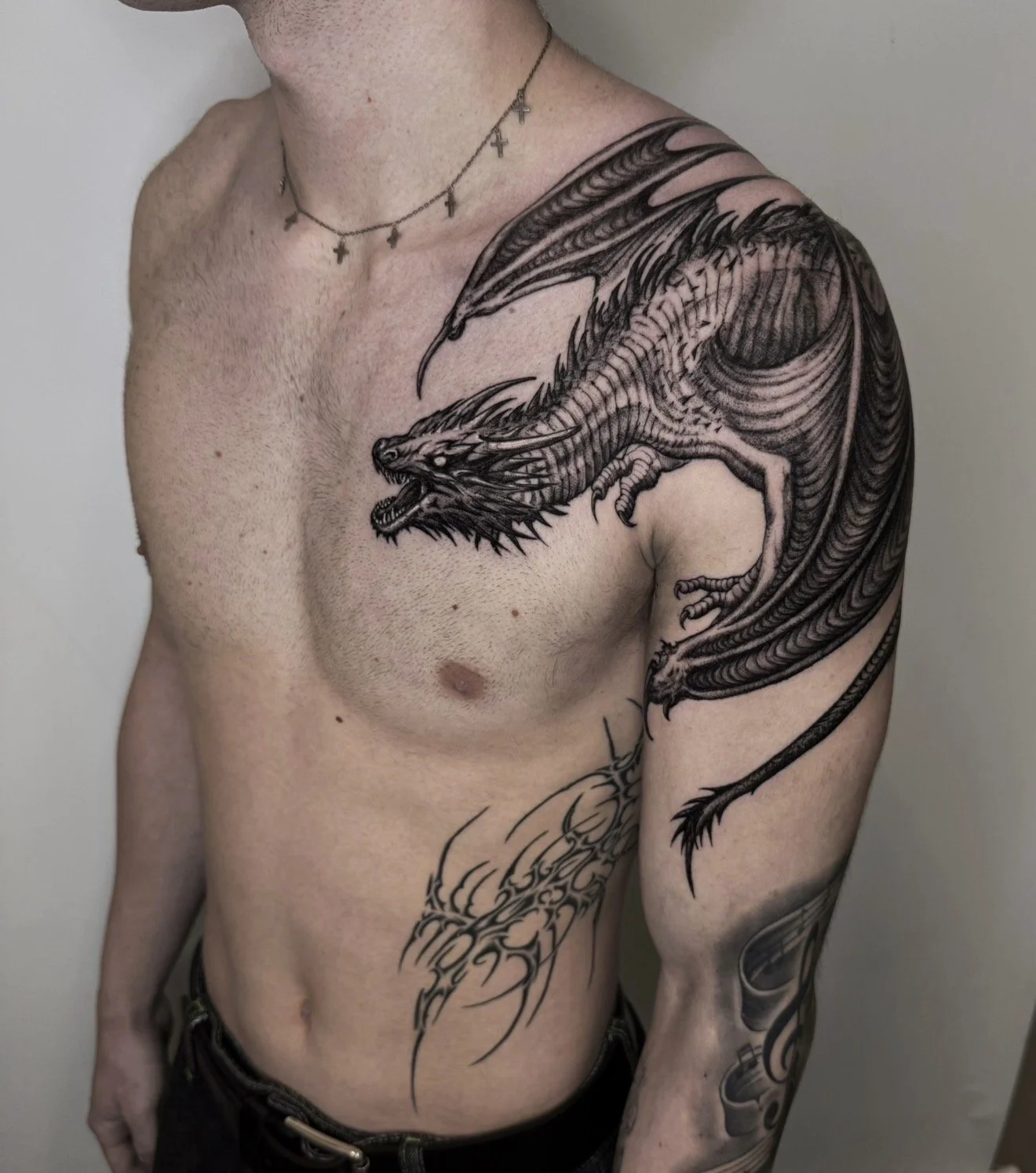 Man with a large black and gray dragon tattoo on his upper chest, shoulder, and arm, and wearing a cross necklace.