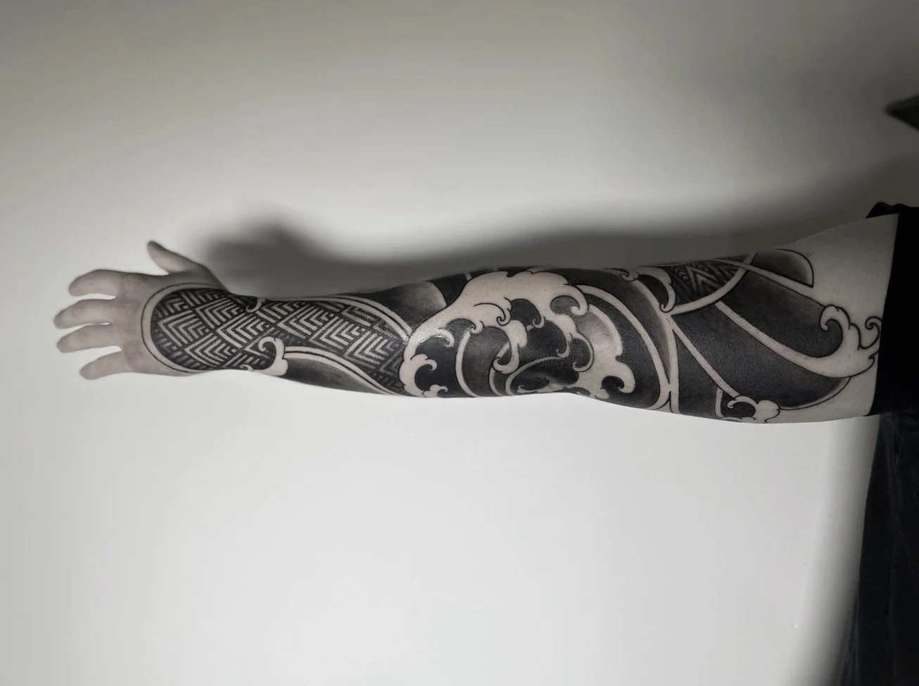 A person's arm with a detailed black and gray tattoo sleeve featuring geometric patterns and wave designs.