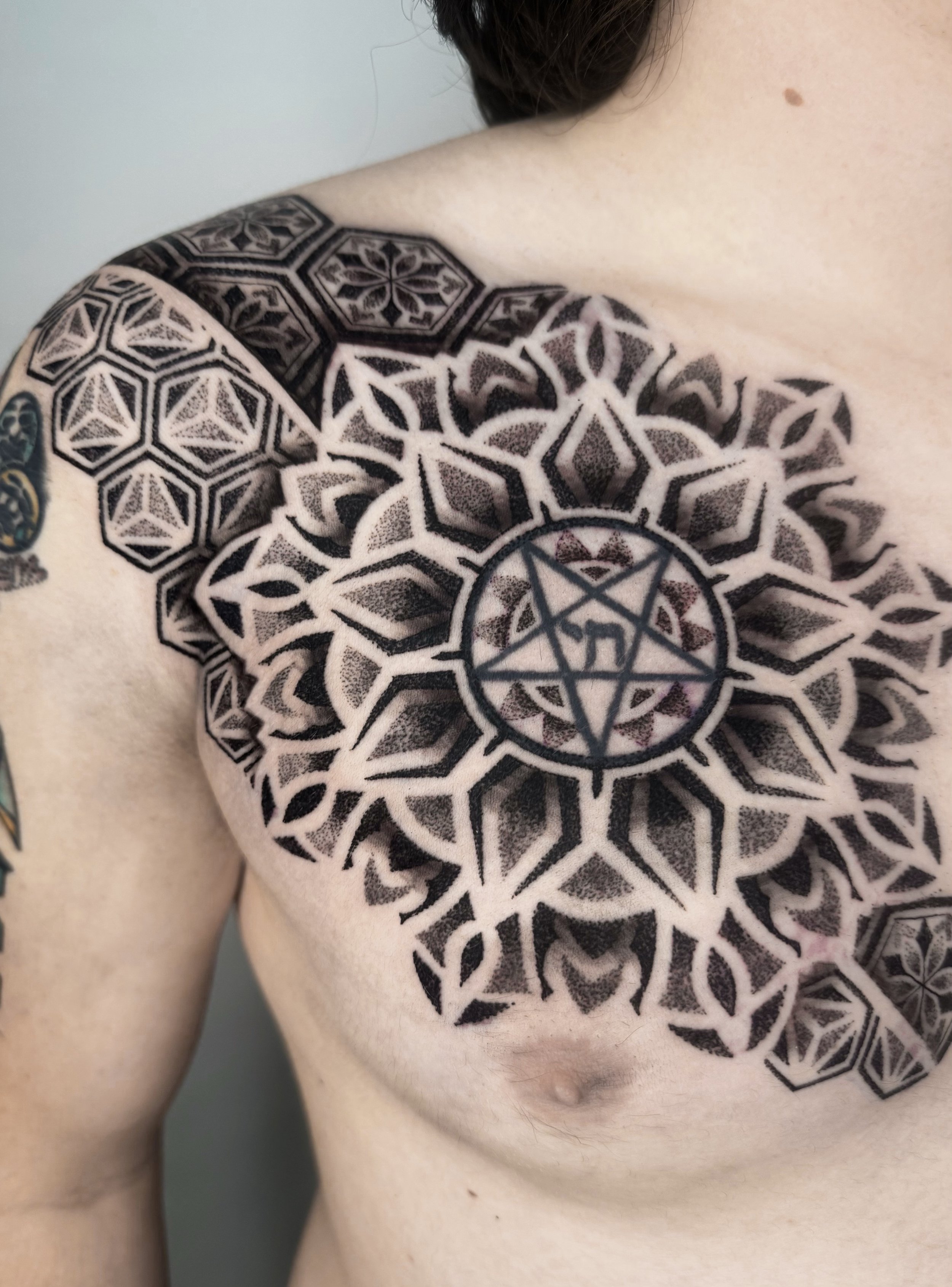 Close-up of a person's shoulder and upper arm with tattoo art featuring geometric patterns and dodecagon shapes, some with floral motifs, and a pentagram enclosed in a circle.