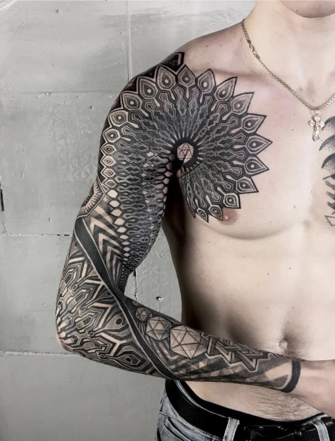 Close-up of a person with extensive black and gray geometric tattoos covering their shoulder, arm, and upper body, wearing a necklace with a cross pendant, standing against a plain concrete wall background.
