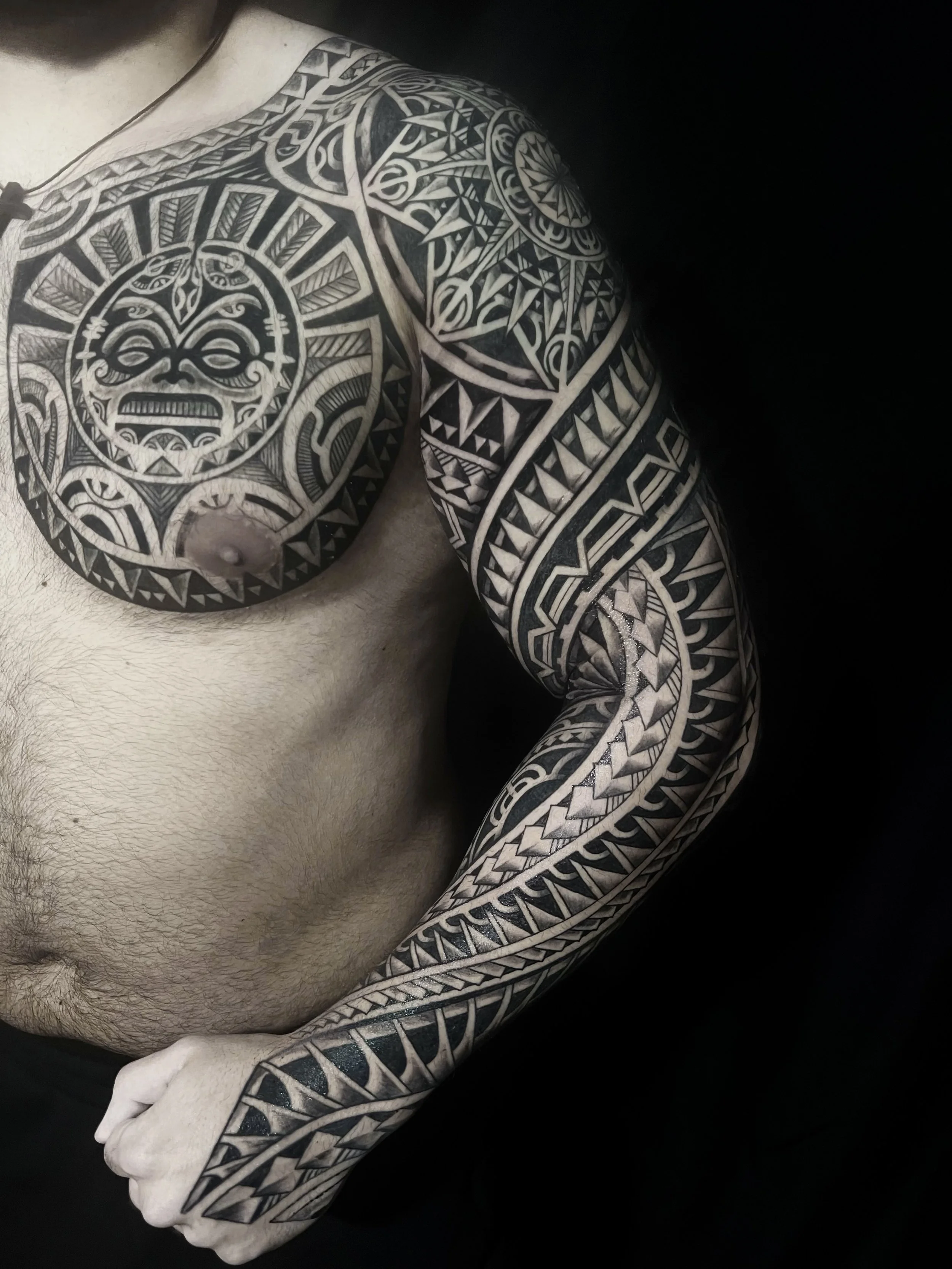 A man with extensive black and gray Polynesian-style tattoos covering his shoulder, arm, and chest, set against a black background.