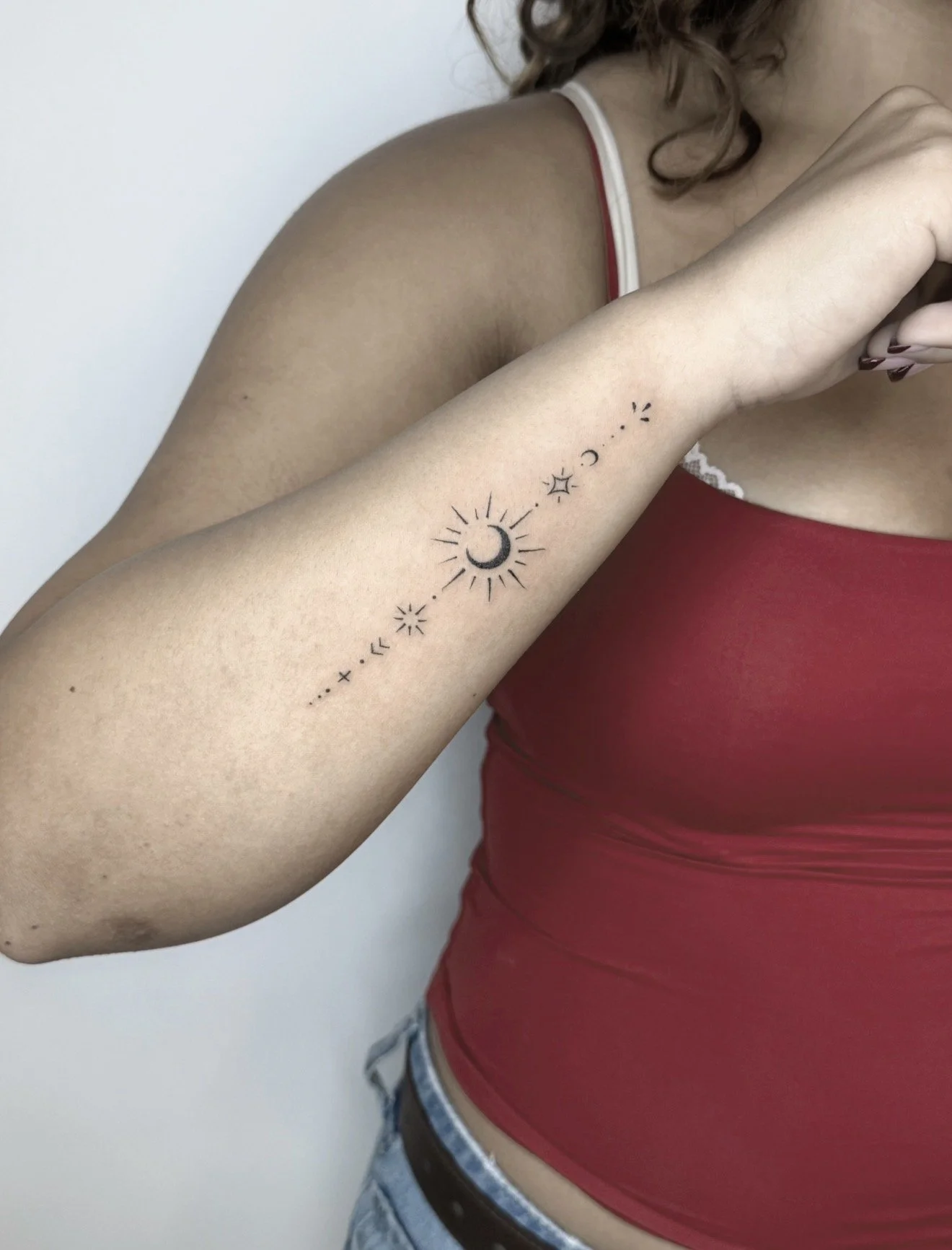 A woman with curly hair, wearing a red sleeveless top and blue jeans, has a moon and star tattoo on her forearm.