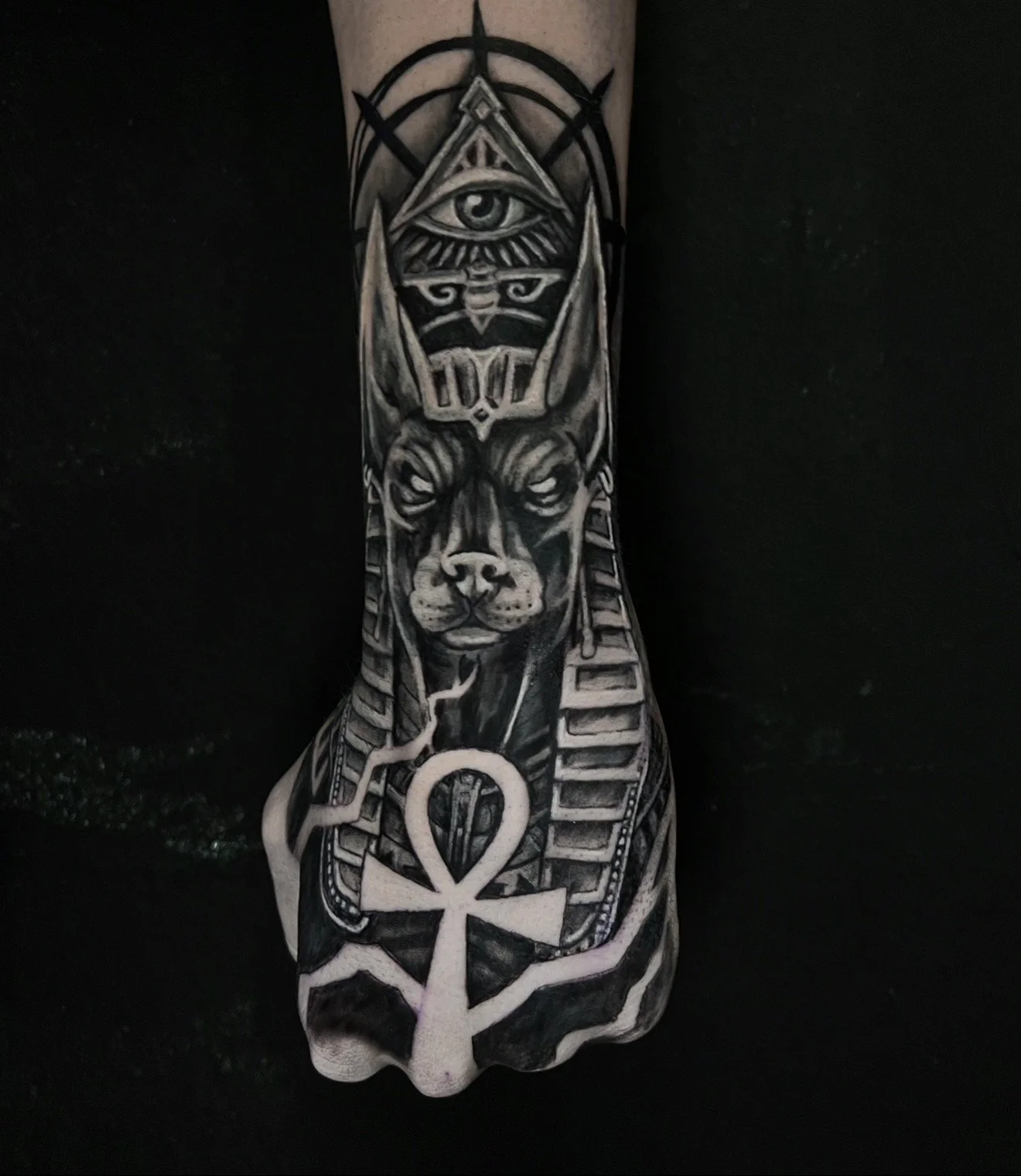 A tattoo on a person's forearm depicting a stylized Egyptian scene with a dog or jackal head in ancient Egyptian style, an eye within a pyramid shape, and various hieroglyphic symbols.