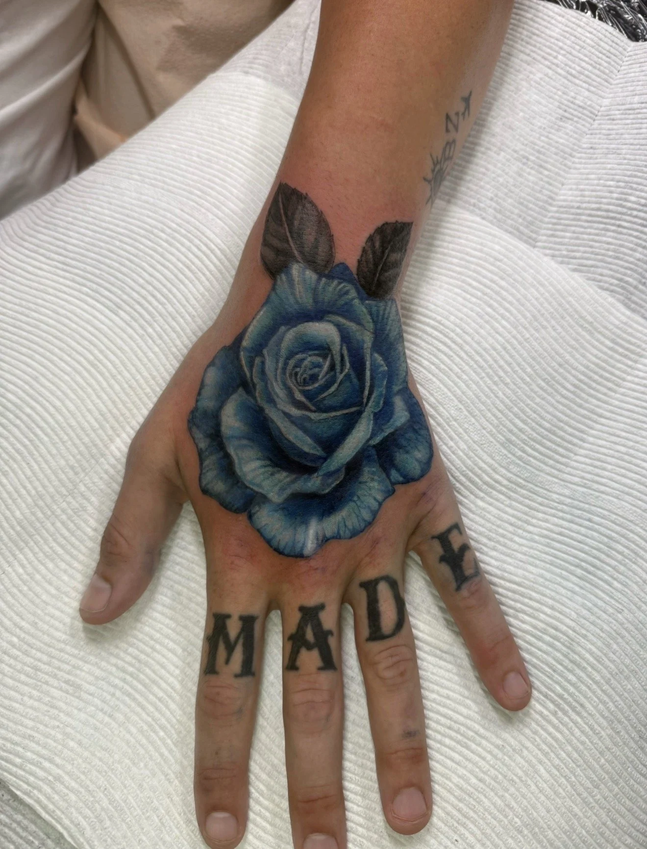 A hand with a large blue rose tattoo on the back of the hand and the word "MAD" tattooed across the knuckles.