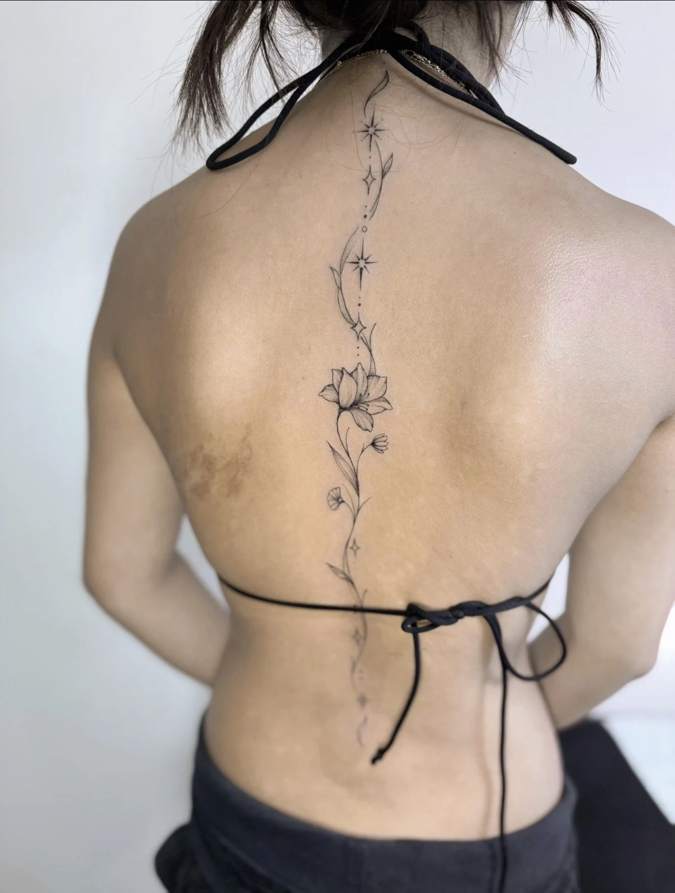 A person with long hair wearing a black backless top, showing a detailed floral tattoo running along their back from the neck to the lower back.