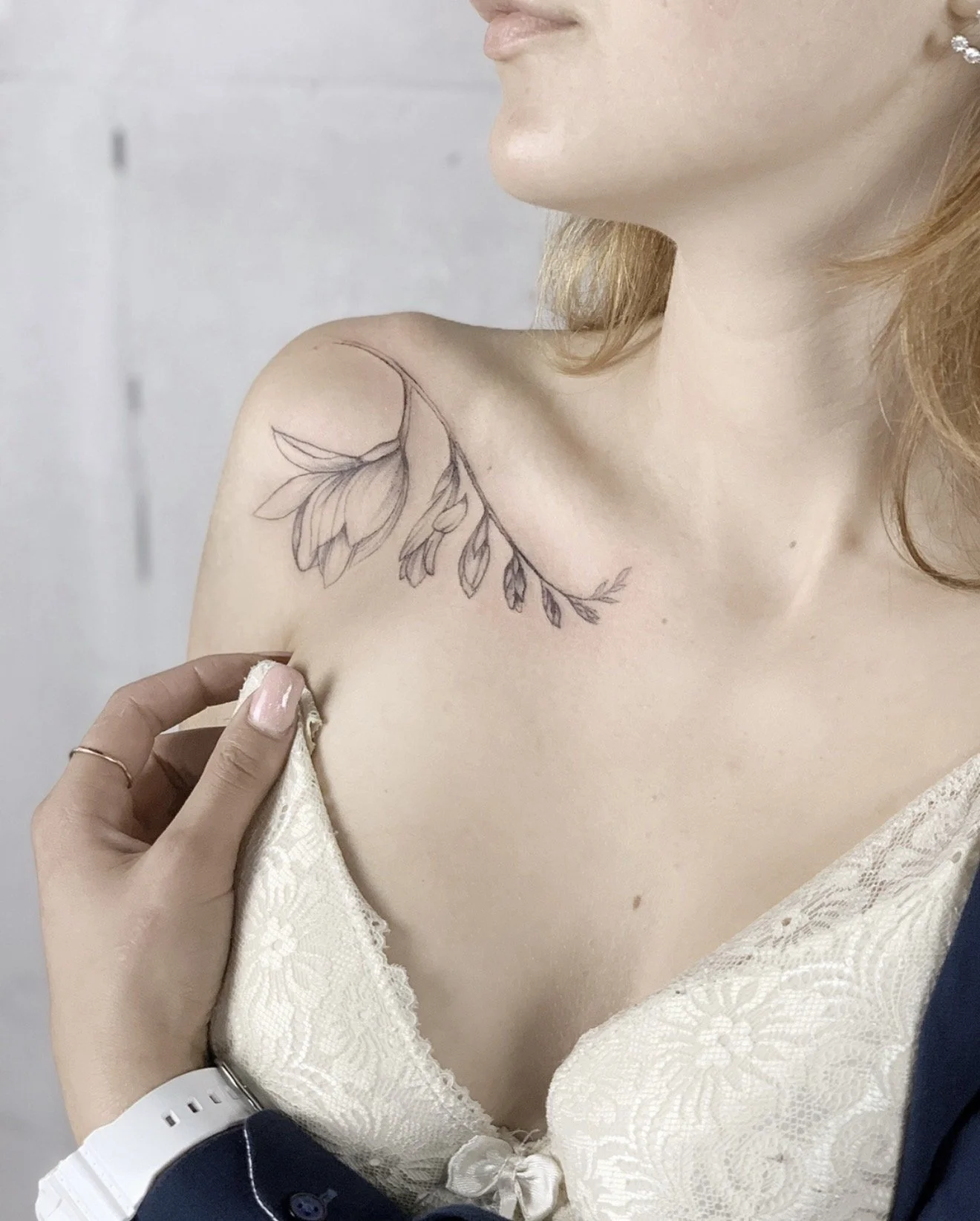 A woman with a floral outline tattoo of leaves and flowers on her shoulder and collarbone area, wearing a white lace top, holding her strap with her left hand, and a person with a dark suit is touching her dress.