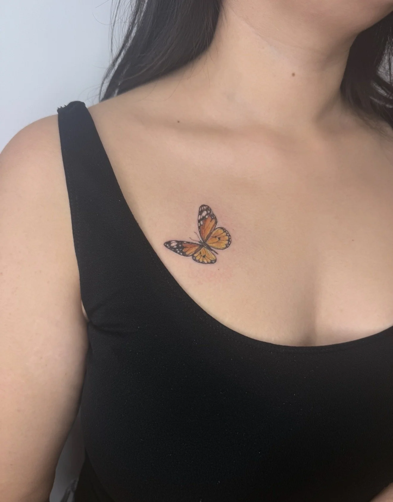 Tattoo of an orange and black butterfly on a woman's upper chest, near the shoulder, wearing a black top.