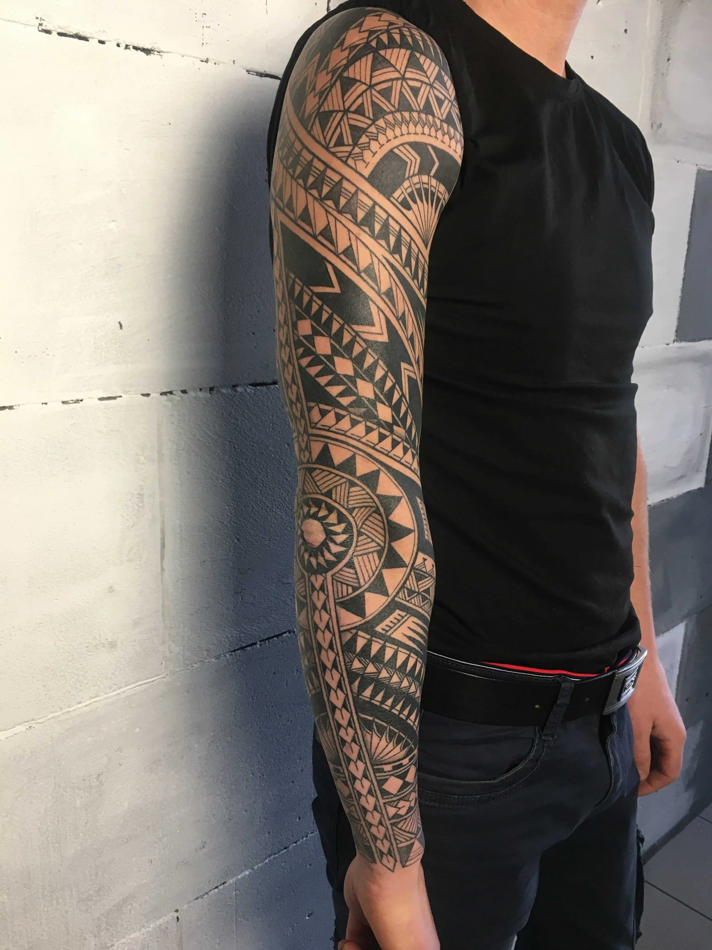 A person with a detailed geometric black tattoo sleeve on their left arm, wearing a black sleeveless shirt and black jeans, standing against a white brick wall.
