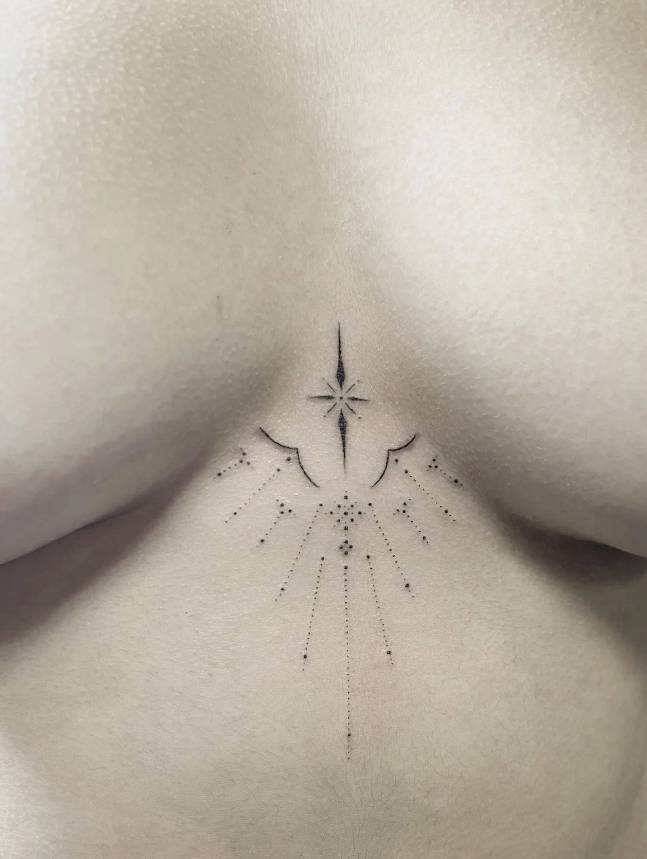 Close-up of a person's lower back and upper buttocks with a black dot tattoo design centered near the waistline, featuring geometric and dotted line elements.