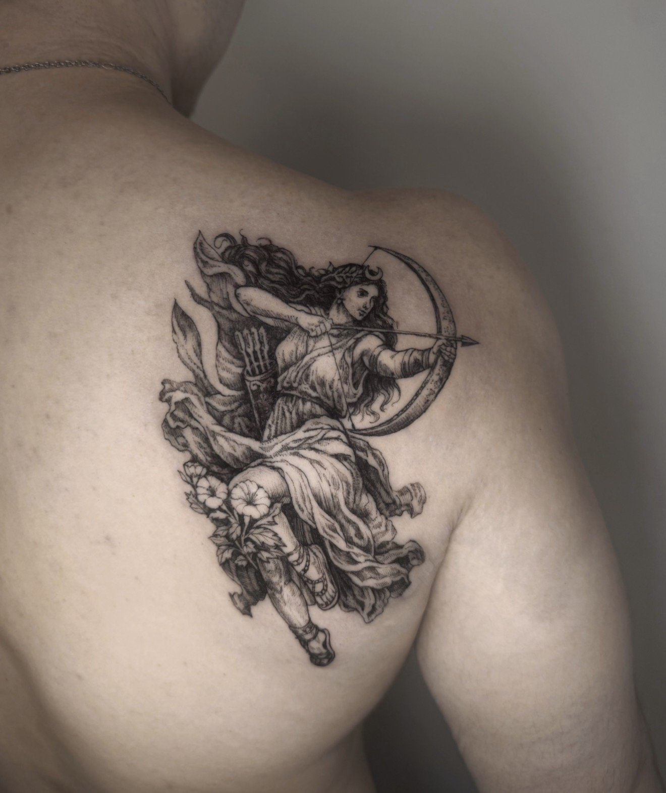 Black and gray tattoo of a detailed female warrior with long hair, holding a bow with an arrow aimed, surrounded by floral and flowing cloth elements on a person's shoulder.