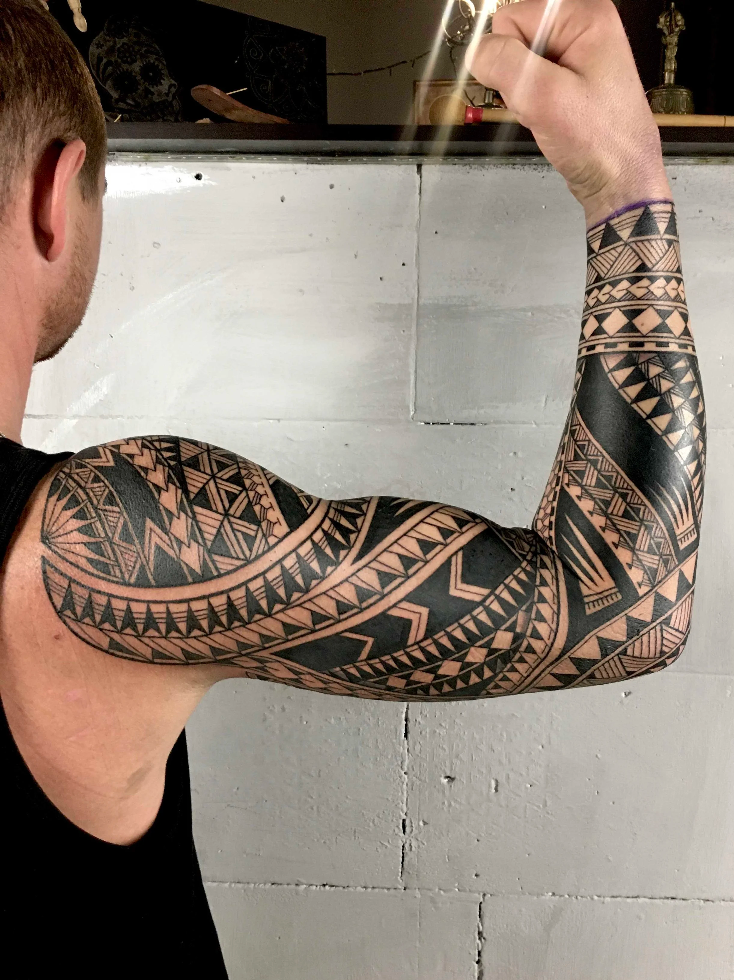 A person with tattoos on their arm flexes their bicep, showing the tattoo design. The person is wearing a black sleeveless top, and a white wall is in the background.