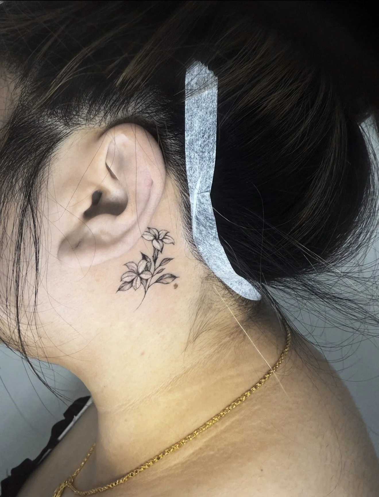 Close-up of a woman's ear and neck, showing a floral tattoo behind her ear on her neck, dark hair with highlights, gold necklaces, and a headband.