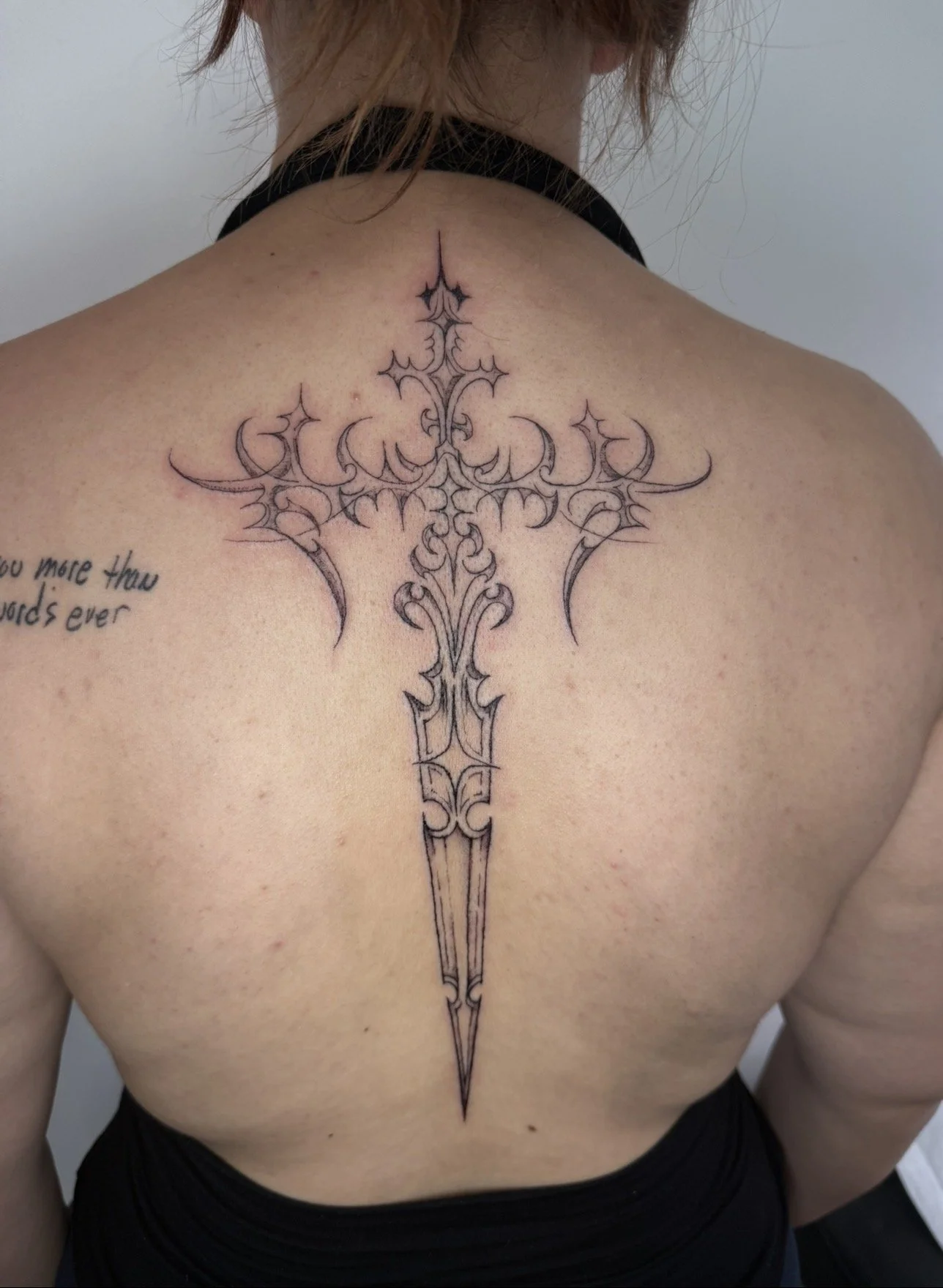 A person with shoulder-length hair and fair skin, showing a detailed black line tattoo of a large cross on their upper back centered along the spine.