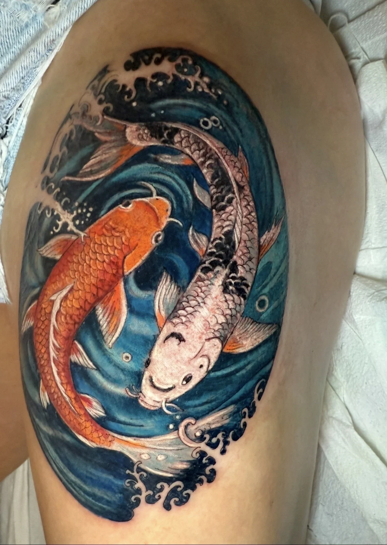 Colorful tattoo depicting two koi fish swimming in a circular pattern, with stylized waves surrounding them.