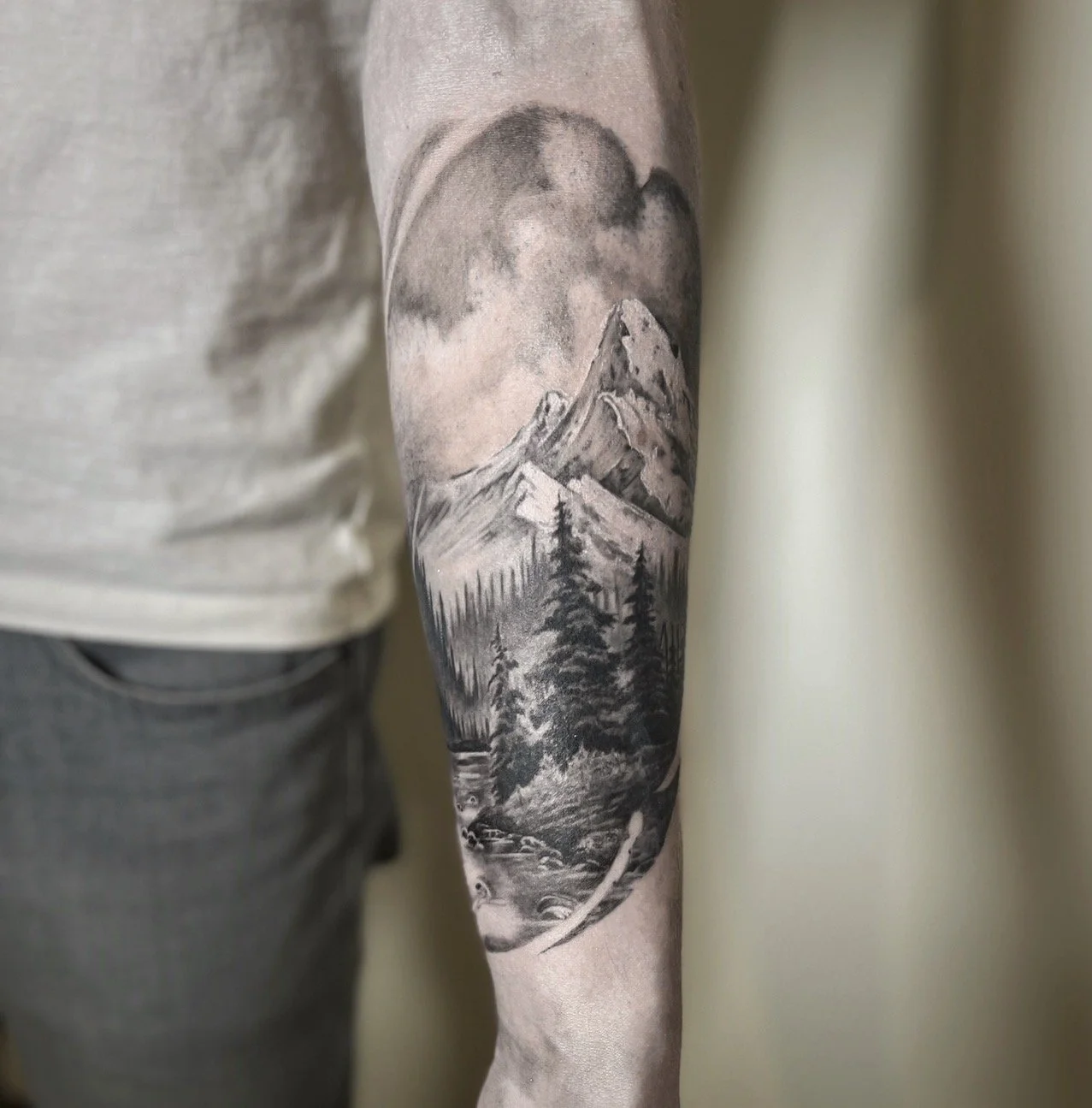 Black and gray mountain landscape tattoo on person's arm showing mountains, trees, and clouds