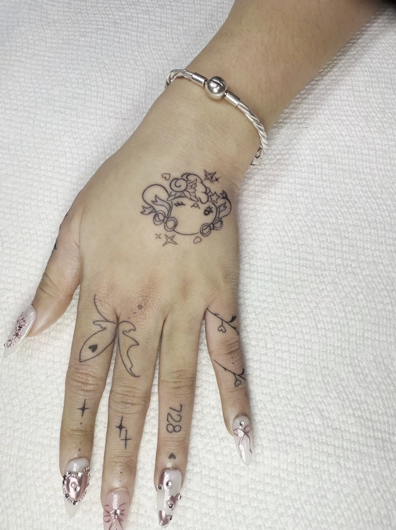 A hand with a silver bracelet, henna tattoos, and long decorated nails, resting on a white textured surface.