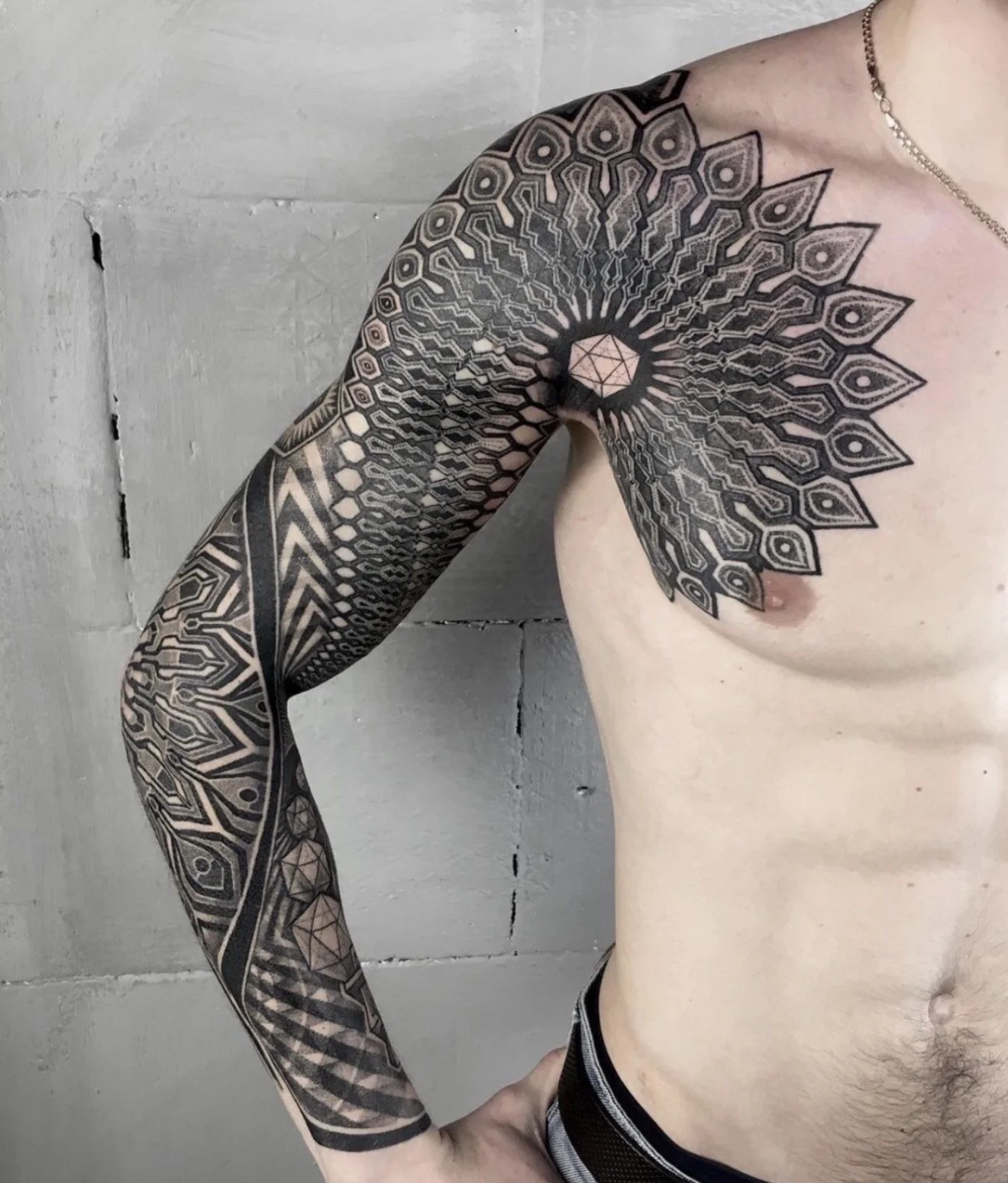 A person with an elaborate black geometric tattoo covering their left shoulder, arm, and chest, standing against a concrete wall.