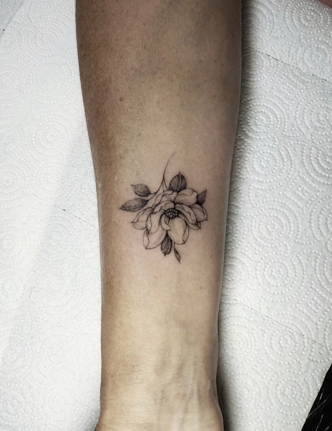 Black ink floral tattoo of a large flower with leaves on the inner forearm resting on a textured paper towel.