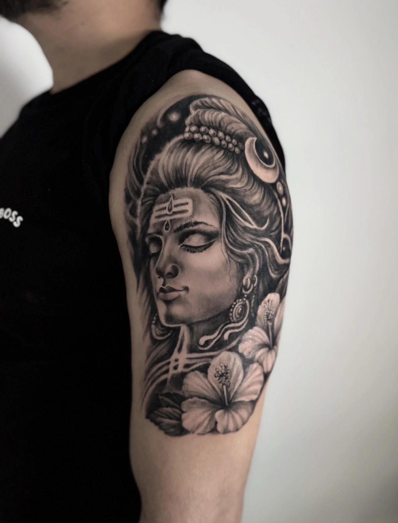 Black and gray tattoo of a serene woman with closed eyes, adorned with jewelry and flowers, on a person's upper arm.