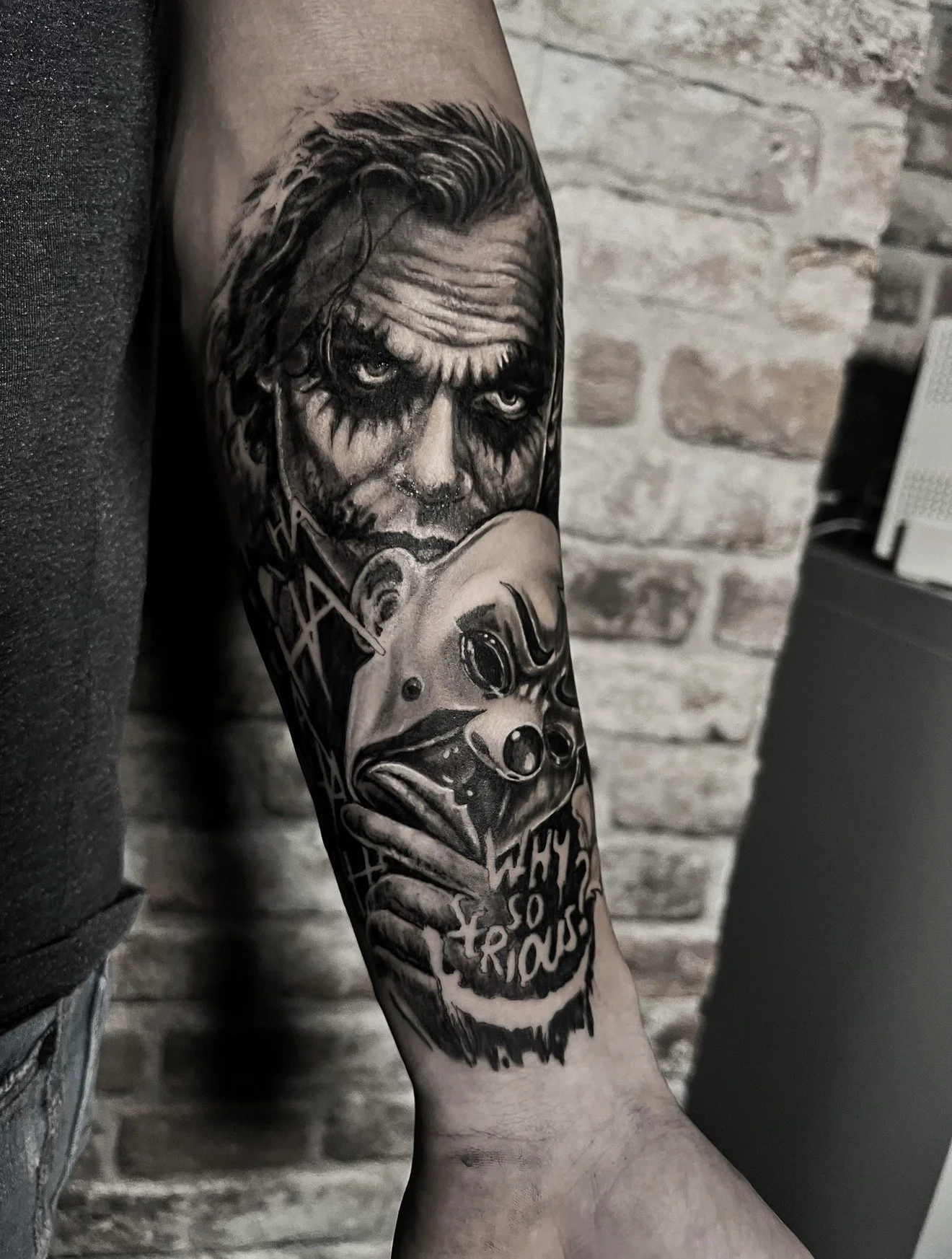 Black and gray tattoo on person's forearm featuring a clown holding a mask with the text 'Why so serious?'