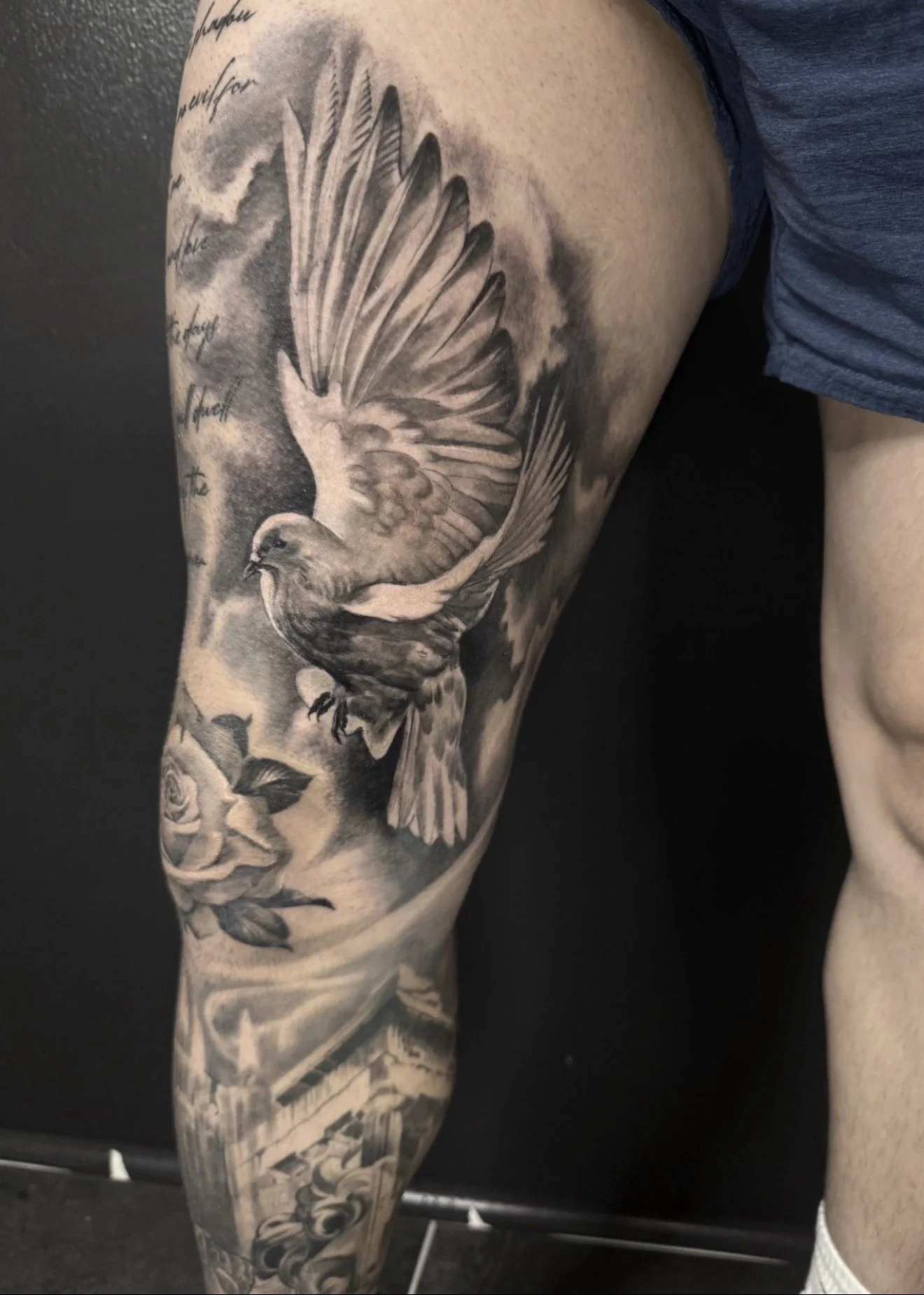 A black and gray tattoo of a bird in flight with outstretched wings on a person's upper arm and shoulder, with some cursive writing on the arm.