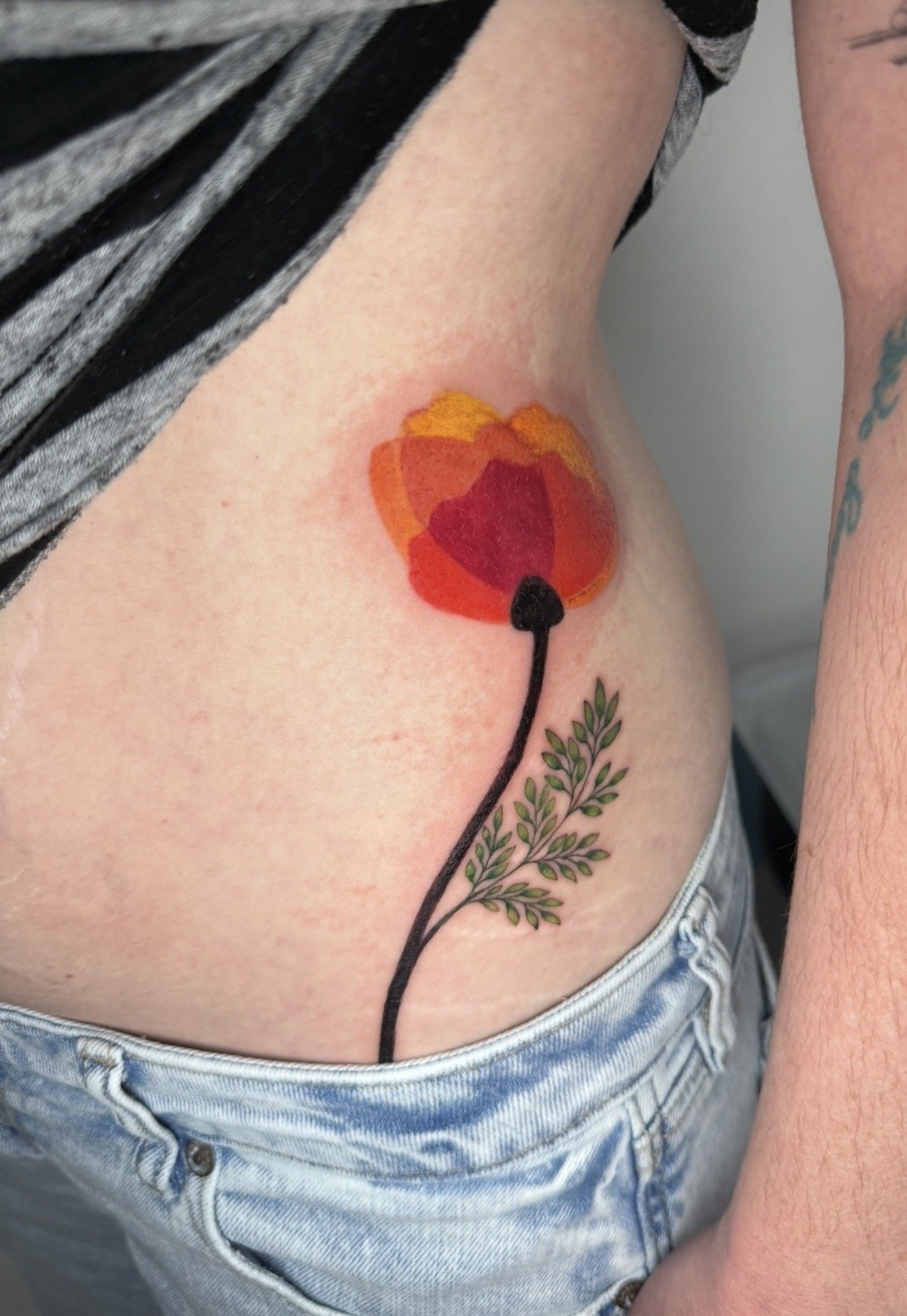 Tattoo of a red-orange flower with a black stem and green leaves on a person's side.