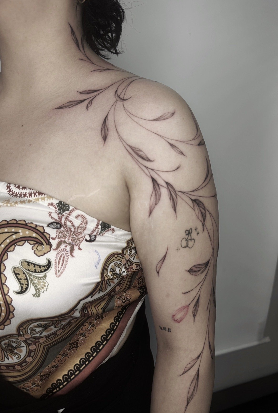 Close-up of a person's shoulder and arm featuring a large floral tattoo with leaves and a small cherry tattoo.