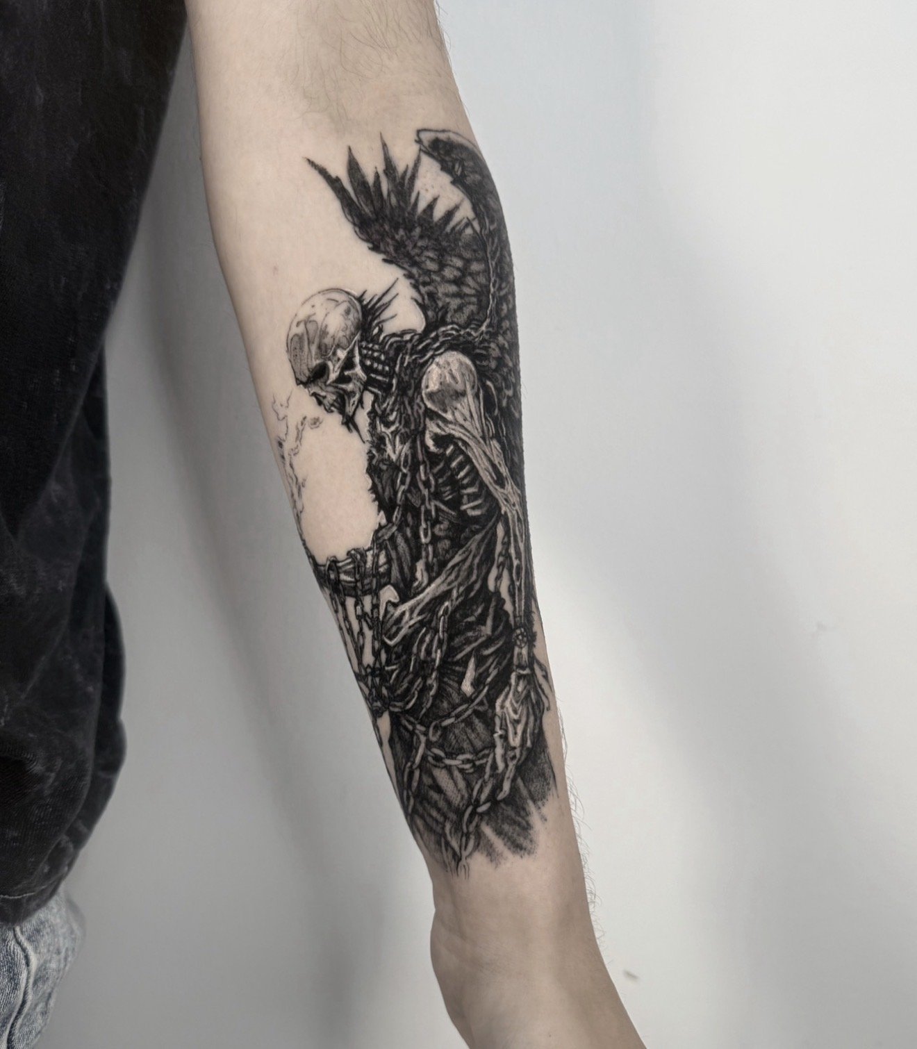 Black and gray tattoo of a skeletal figure with wings, chains, and dark shading on a person's forearm.