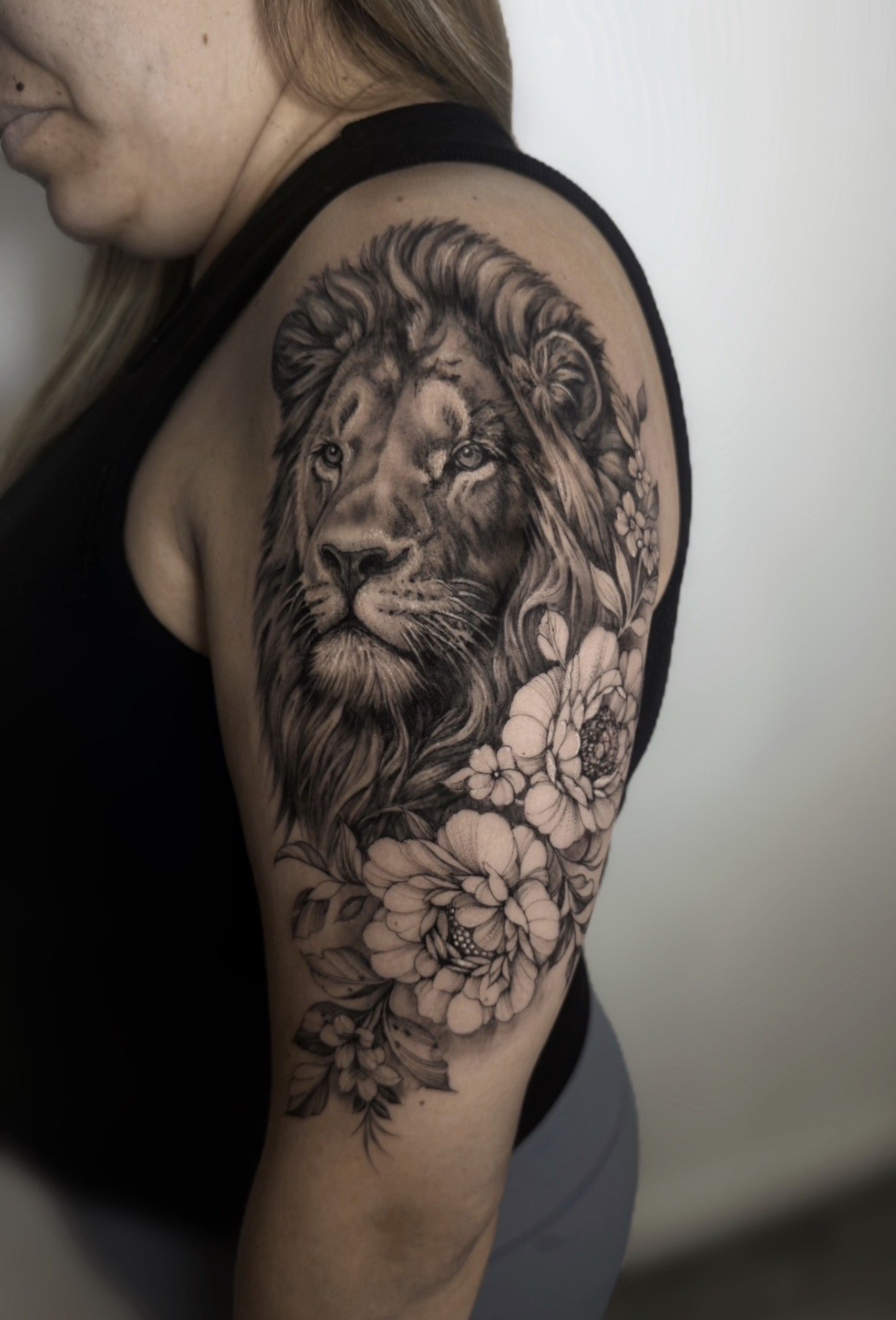 Black and gray tattoo of a lion's face with flowers on the arm of a woman with light skin, wearing a black sleeveless top.