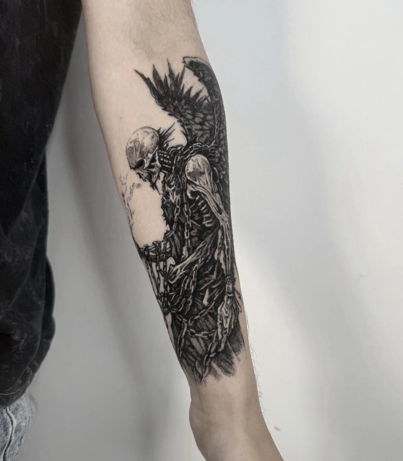 Black and gray tattoo of a skeletal figure with large wings, chains, and a broken sword on a person's forearm.