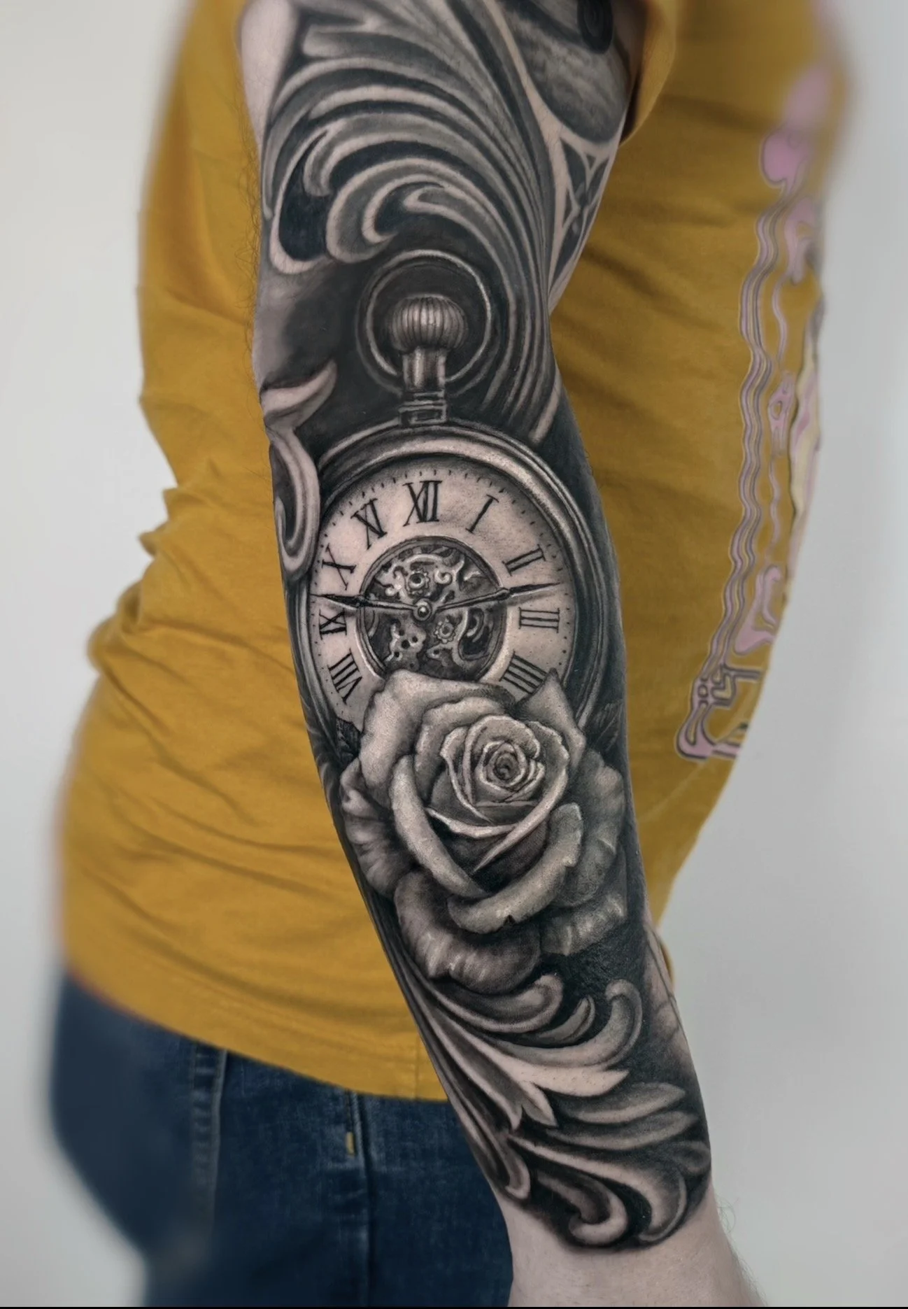 Tattoo sleeve depicting a vintage pocket watch, roses, and swirling patterns on a person's arm, with a yellow shirt and blue jeans.
