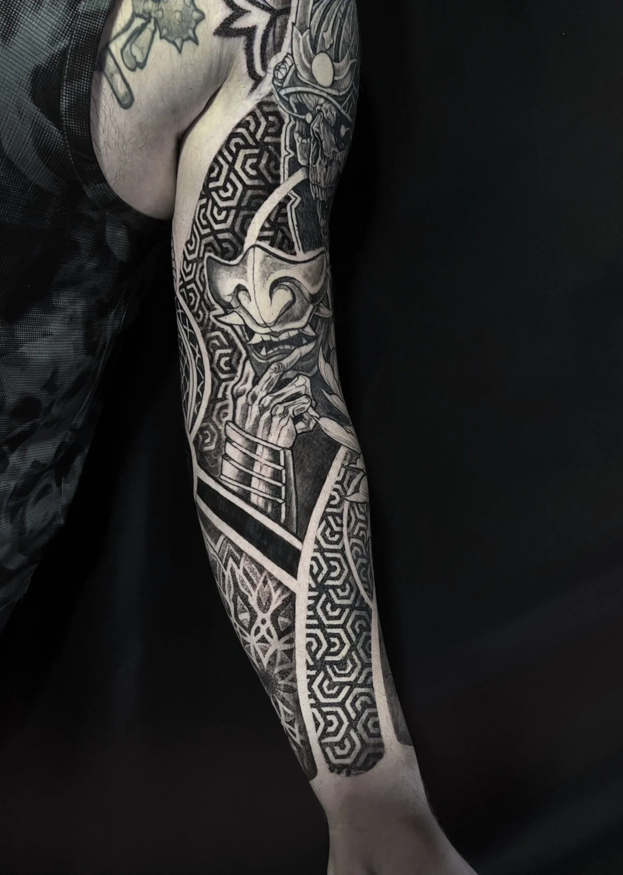 A person's arm with a detailed black and gray tattoo of a traditional Japanese demon mask, geometric patterns, and floral designs.